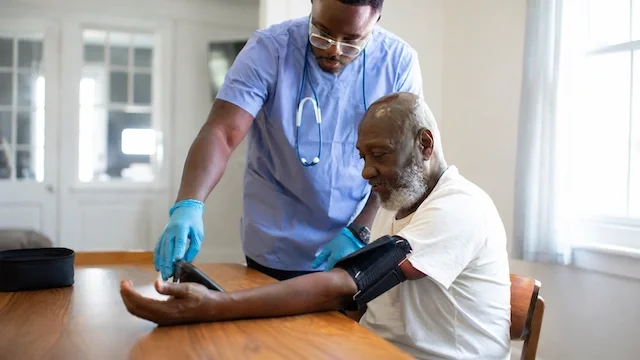 A healthcare professional taking a patient’s blood pressure at a wooden table in a brightly lit room.