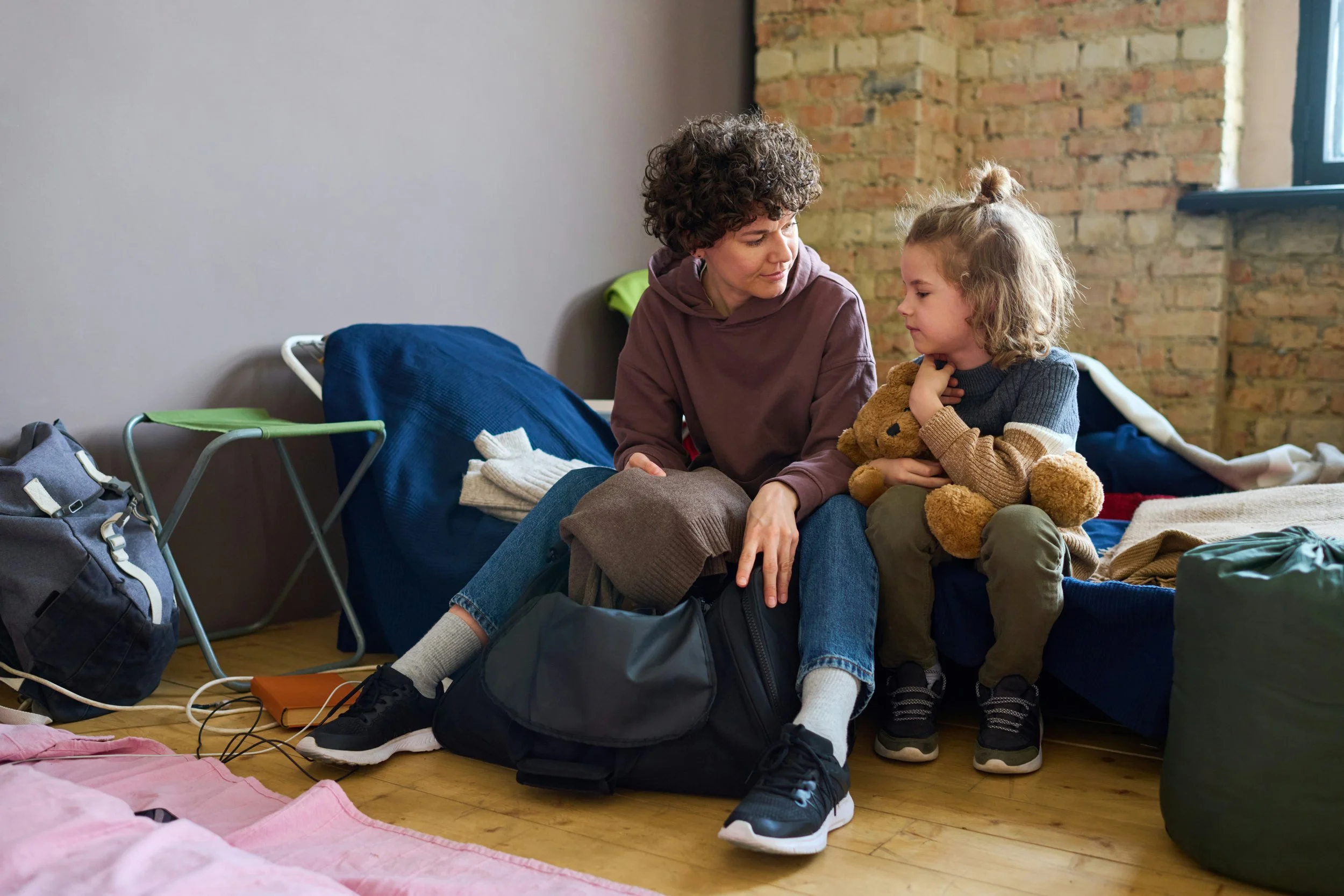 A woman and a child sitting on a bed in a room with a brick wall and a window, surrounded by luggage and clothing.