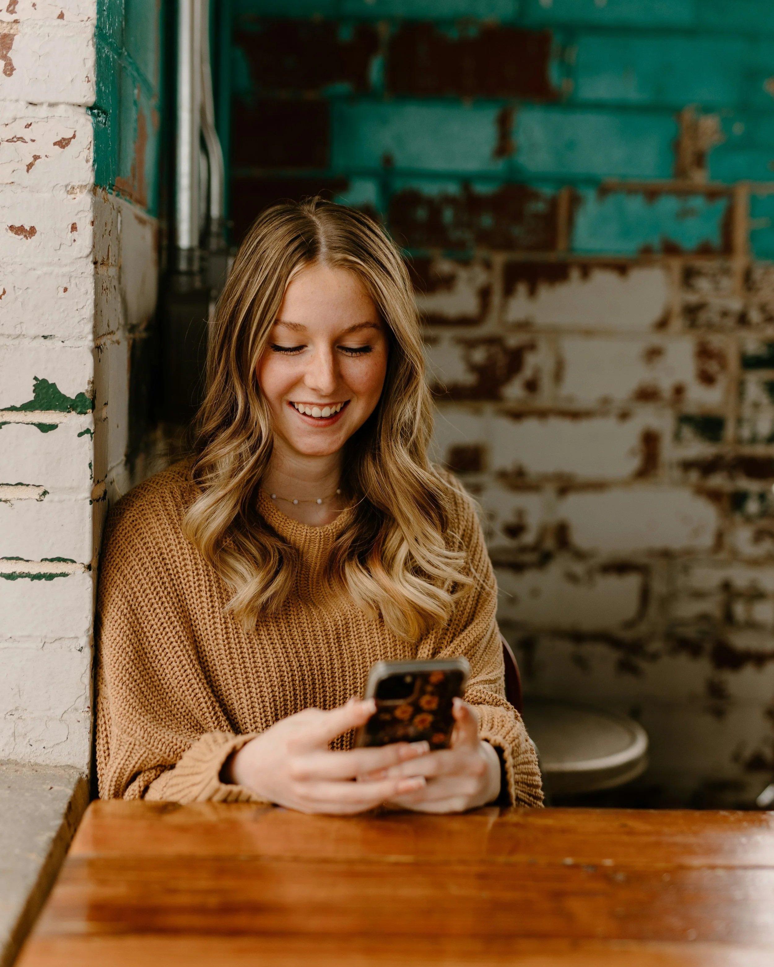 A young woman with blonde hair, wearing a brown sweater, sitting at a wooden table in a rustic cafe, looking at her smartphone and smiling.