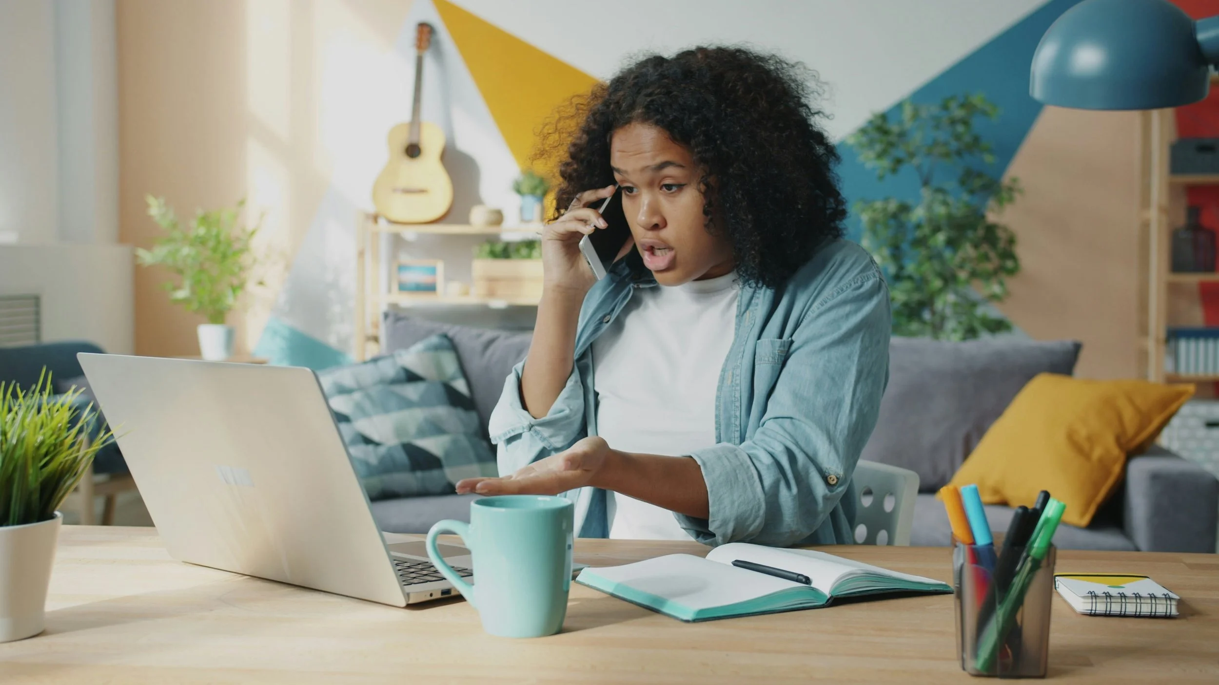 A woman with curly hair talking on her cellphone at a desk with a laptop, notebook, mug, and pens in a colorful living room.