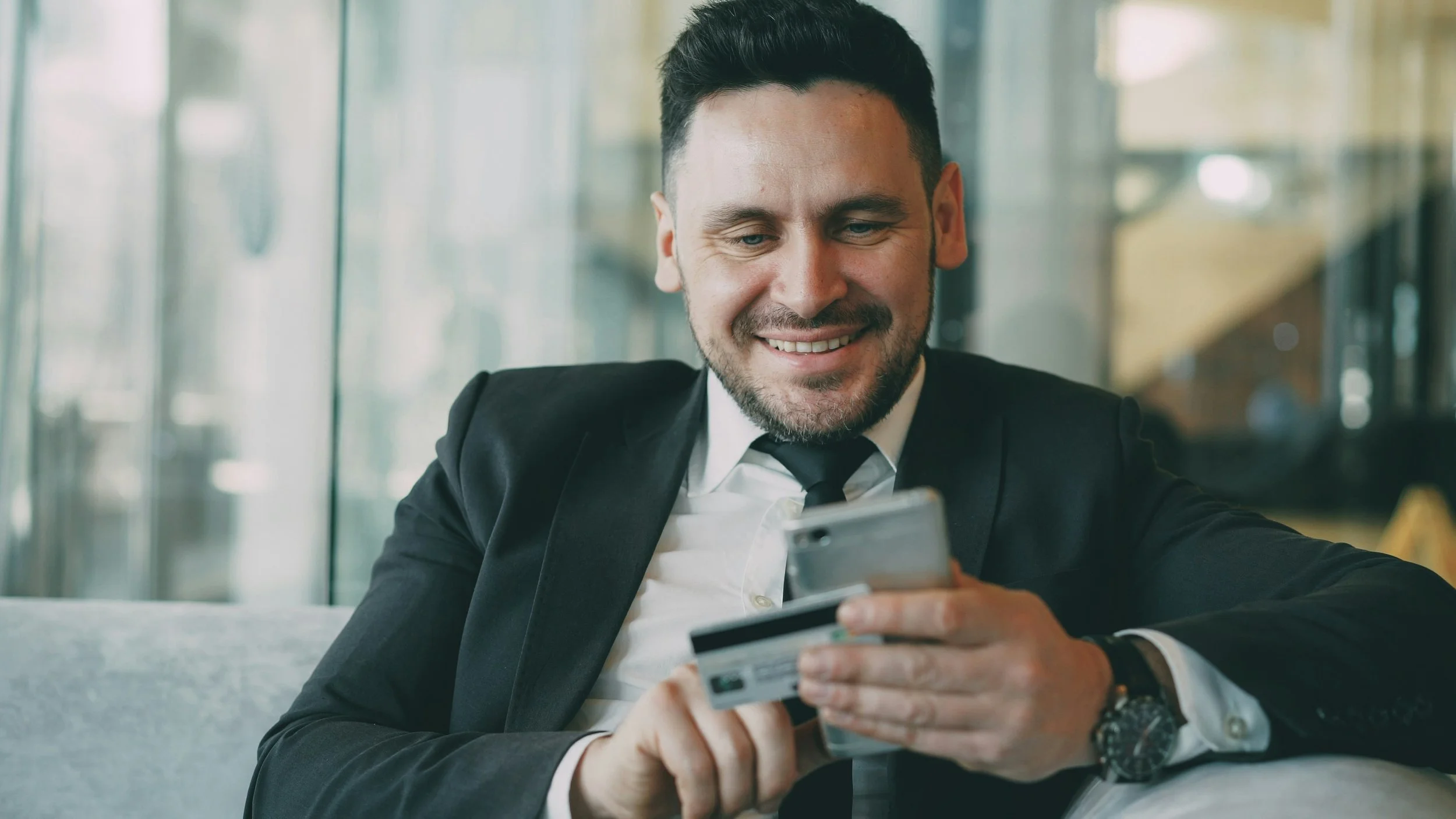 A man in a black suit with a white shirt and black tie, sitting at a table in a modern indoor setting, smiling as he looks at his smartphone and holding a credit card.
