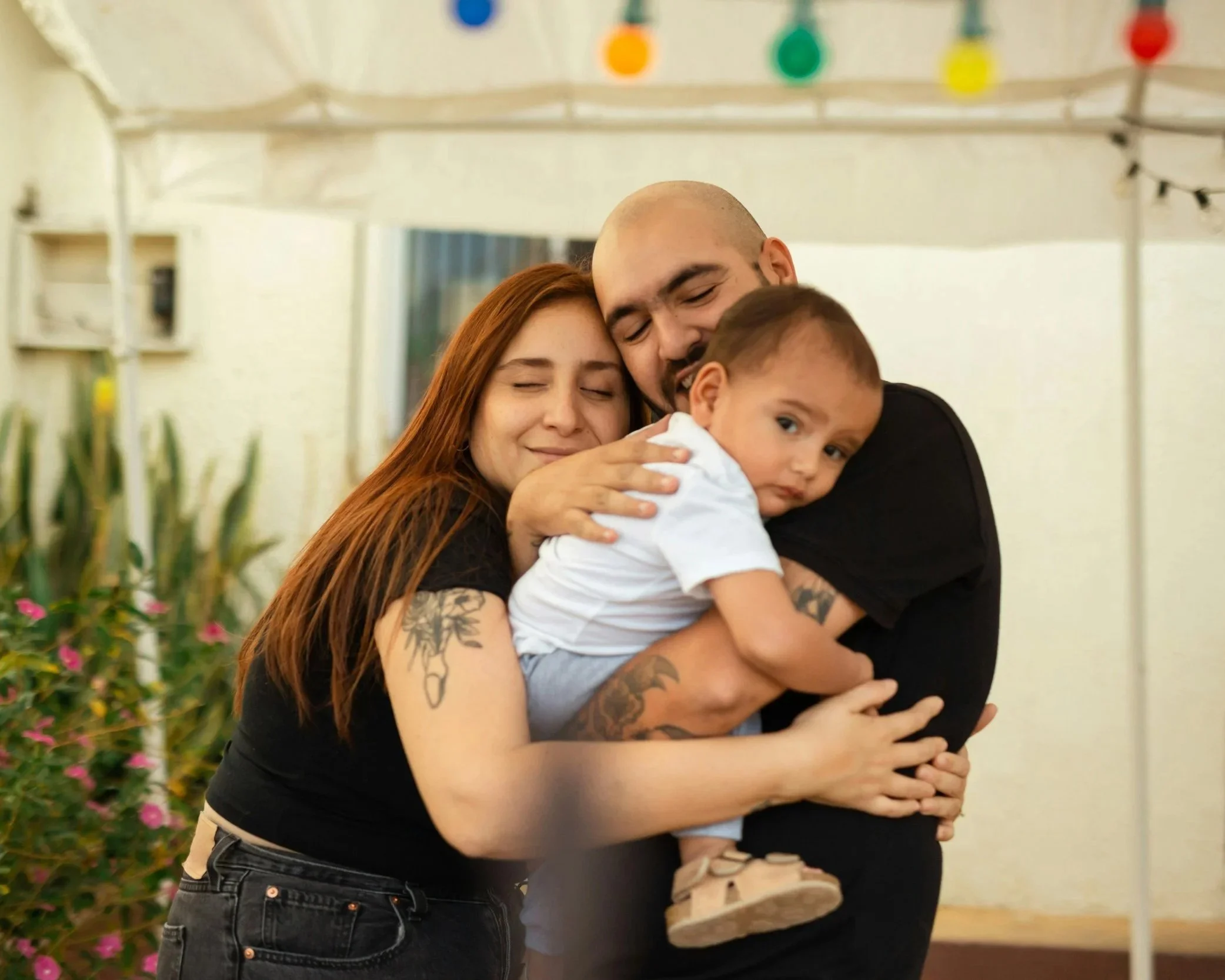 A family of three hugging inside a decorated tent, with a woman with long red hair and tattoos, a man with a shaved head, and a young boy with short hair.