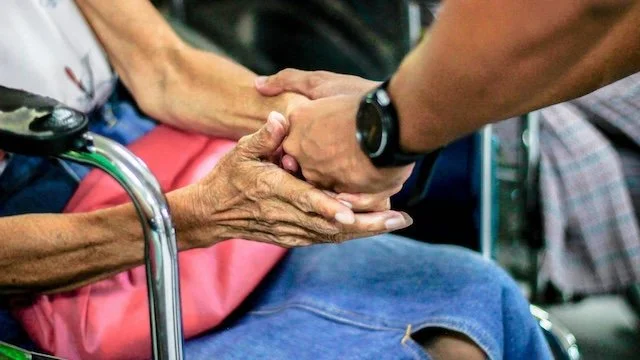 A person in a wheelchair holding hands with an elderly person, providing comfort and support.
