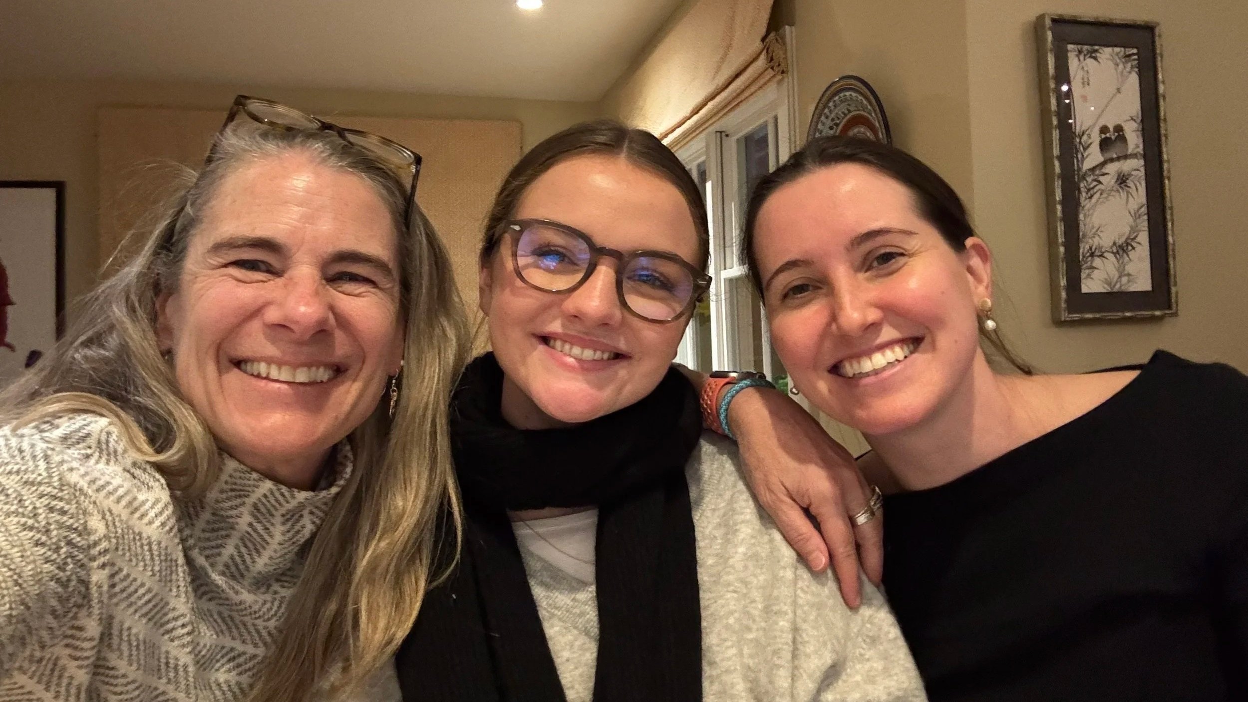 Three women smiling and posing closely together indoors, with pictures and decorations on the wall behind them.