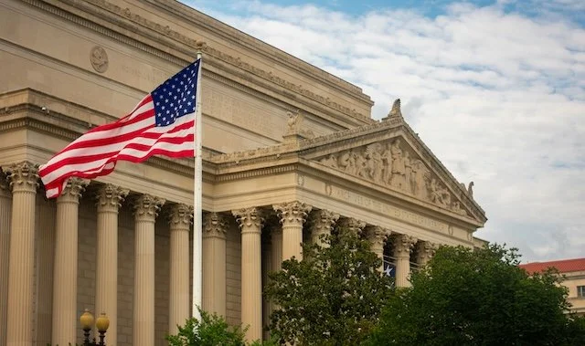 American flag flying in front of a neoclassical building with tall columns and detailed stone carvings, under a partly cloudy sky.