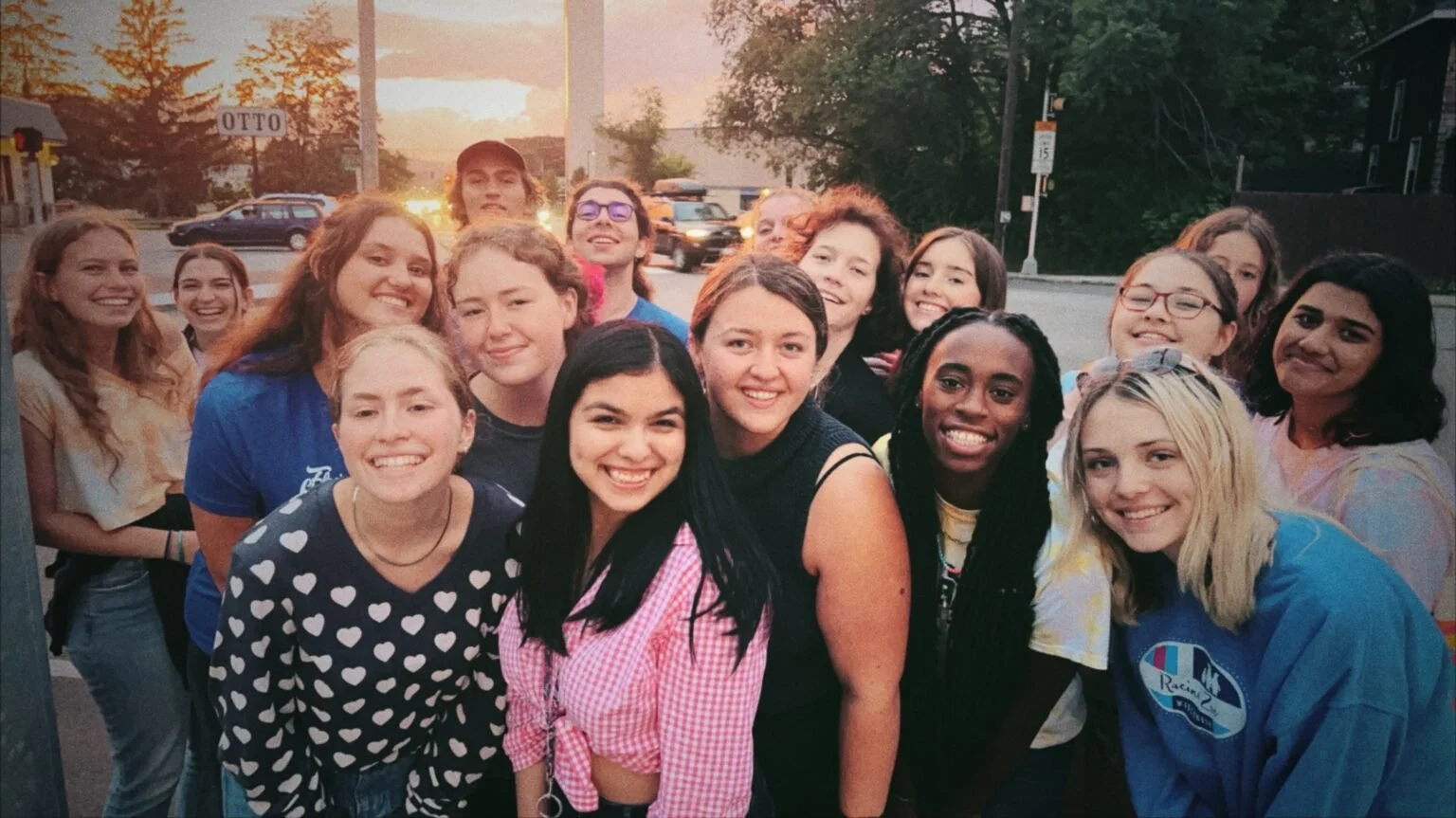 A group of young women and girls smiling together outdoors during sunset, posing for a photo on a city street.