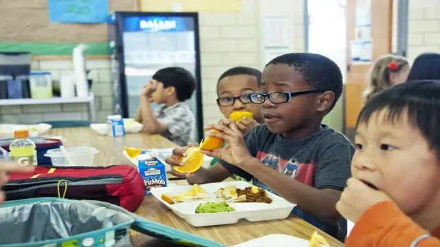 Kids sitting at a cafeteria table eating lunch, with food trays, juice boxes, and a vending machine in the background.