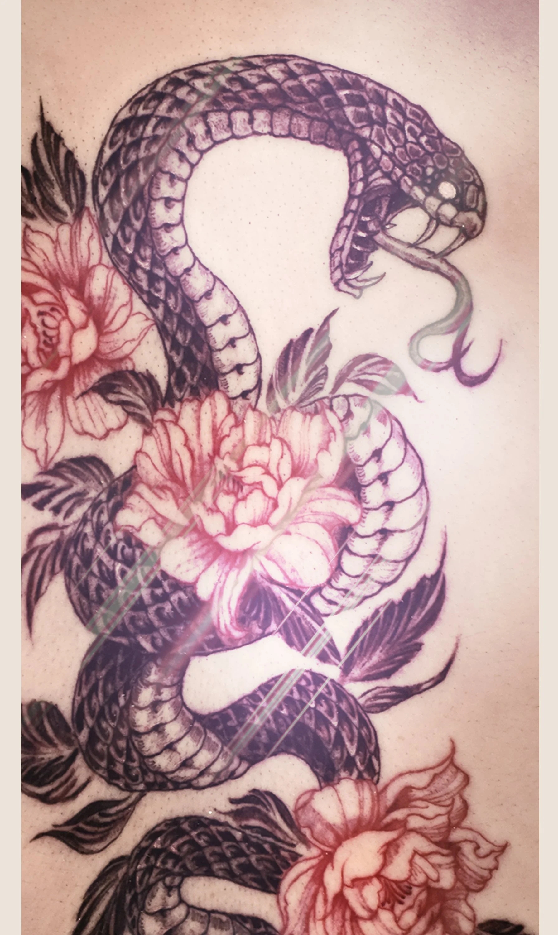 A tattoo of a snake intertwined with flowers and leaves. The snake is coiled around the flowers, appears to be biting its own tail, and has detailed scales. The flowers have large petals and the overall style is black and gray with pinkish accents.