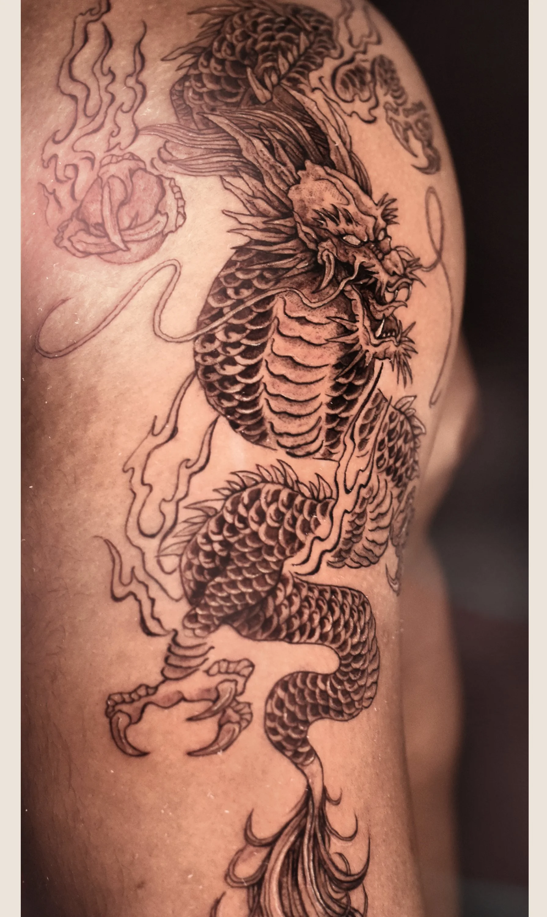 Black outline tattoo of a fierce Chinese dragon with scales, horns, whiskers, and surrounded by clouds, on person's arm.
