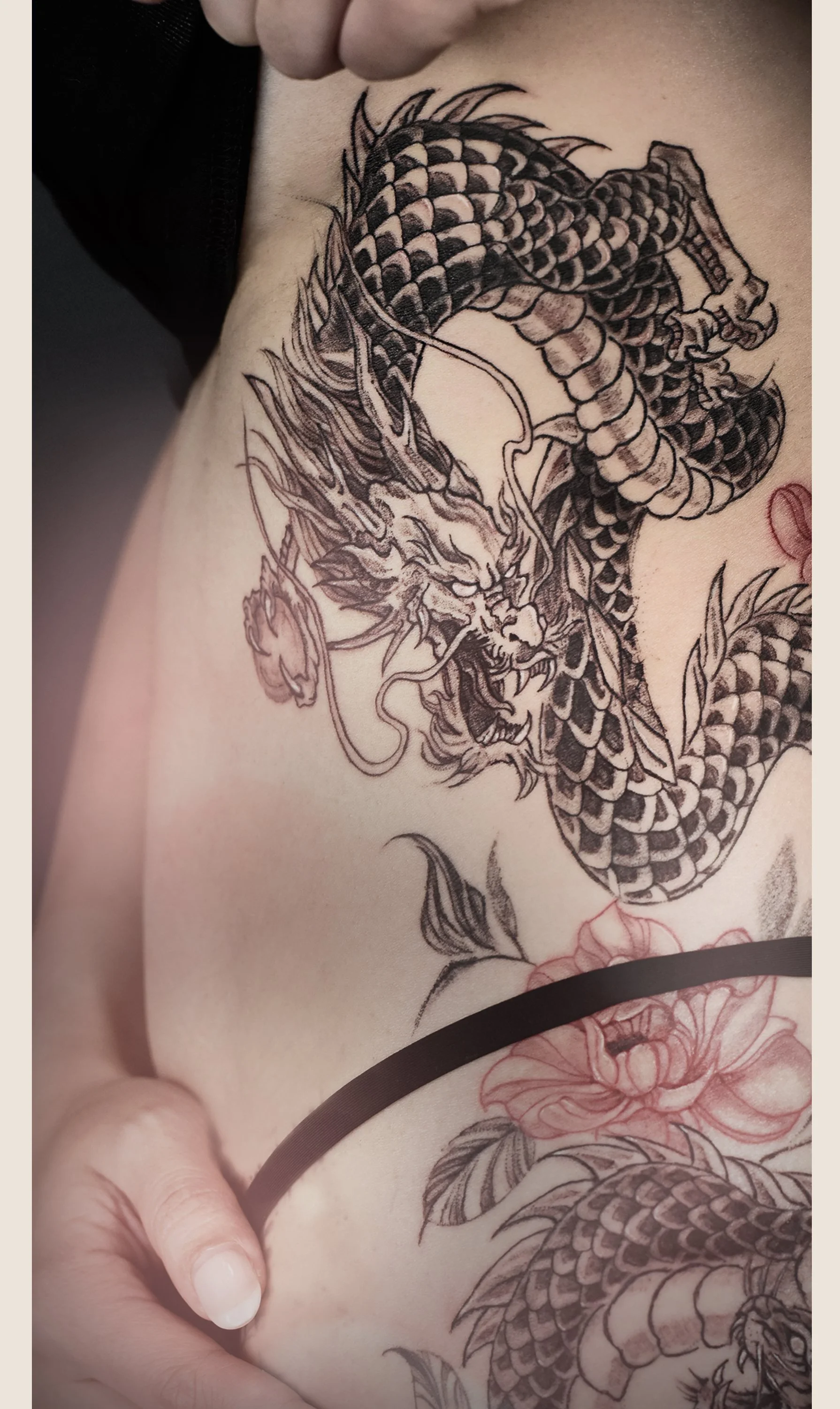 Tattoo of a detailed, fierce dragon with scales, horns, and claws on a person's chest and side, accompanied by a pink flower.