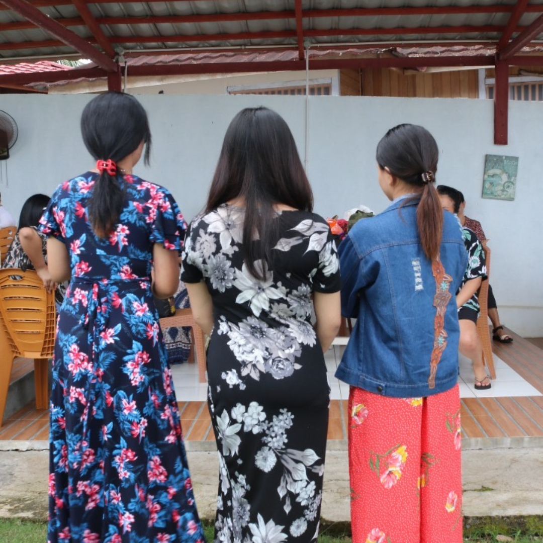 Three women wearing floral dresses and a denim jacket with long hair gather outdoors near a table with flowers, while other people are seated in the background.