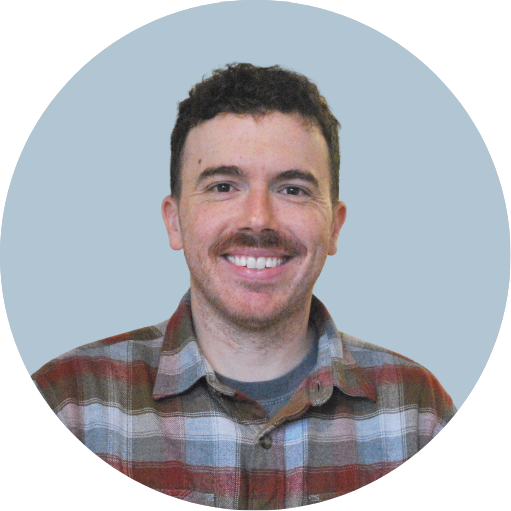 A man with short curly brown hair, a mustache, and a beard smiling, wearing a plaid shirt, against a light teal background.