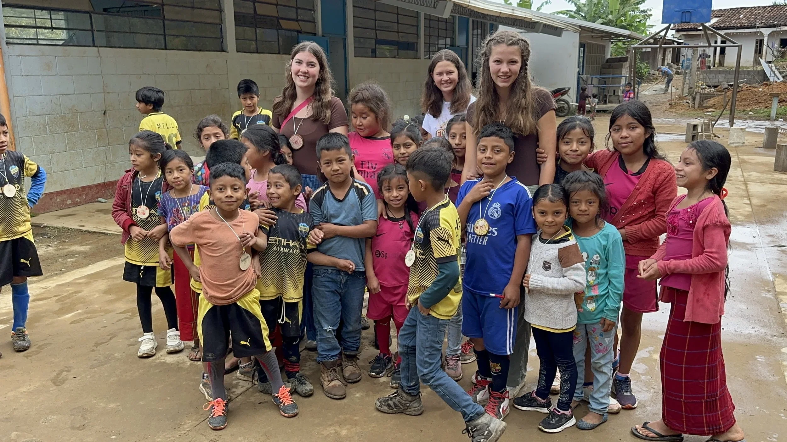 A group of children and four young women standing outdoors in a muddy area, smiling for the camera. The children wear casual clothes and some have medals around their necks. The background shows a cement wall, some construction, and a few other children playing.