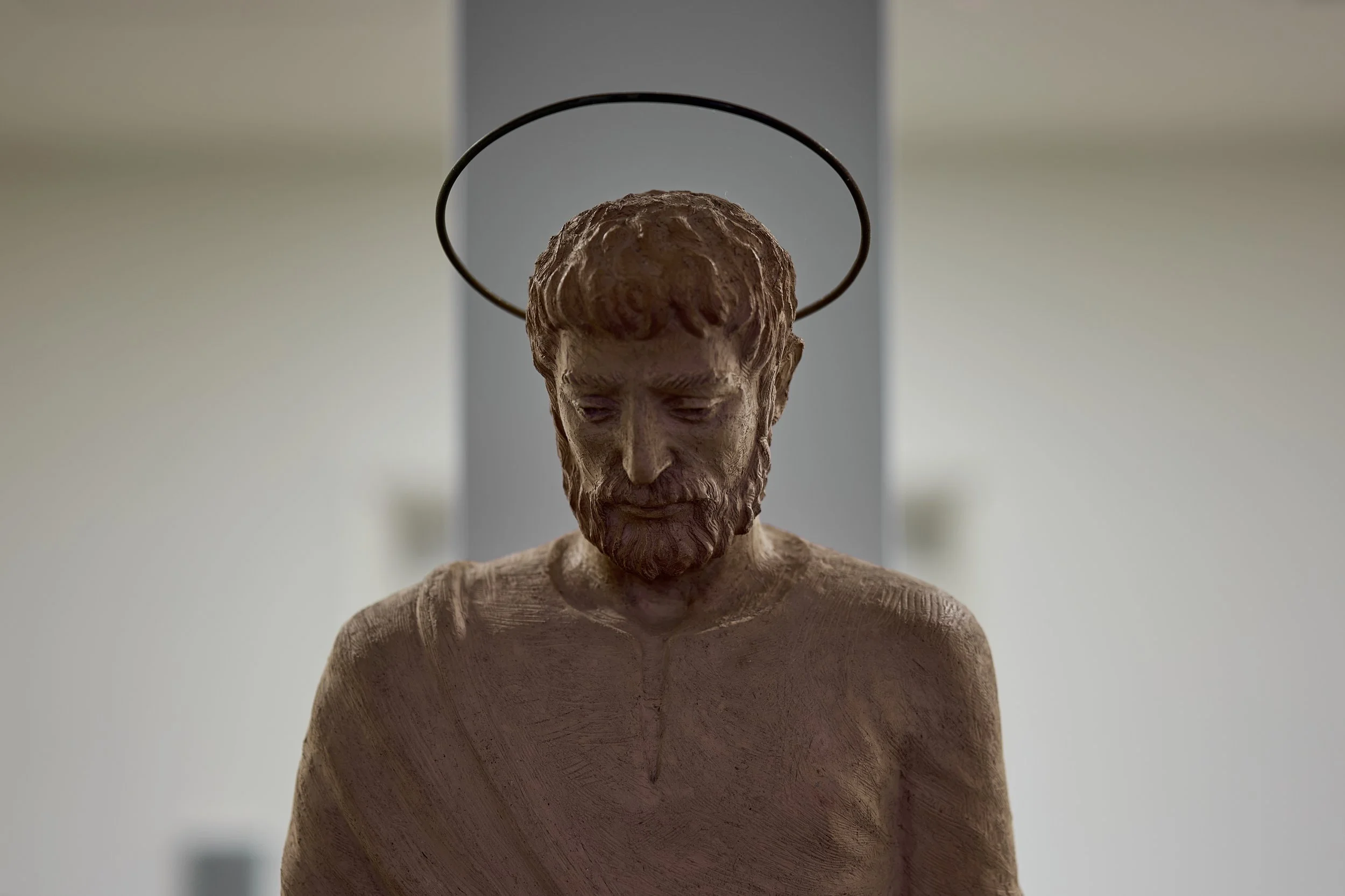 Close-up of a carved statue of Jesus Christ with a halo, looking downward, against a blurred background.