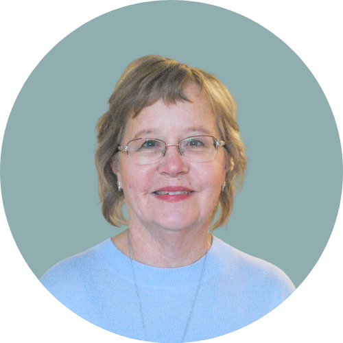 A smiling middle-aged woman with short light brown hair, glasses, and earrings, wearing a light blue shirt, against a teal background.