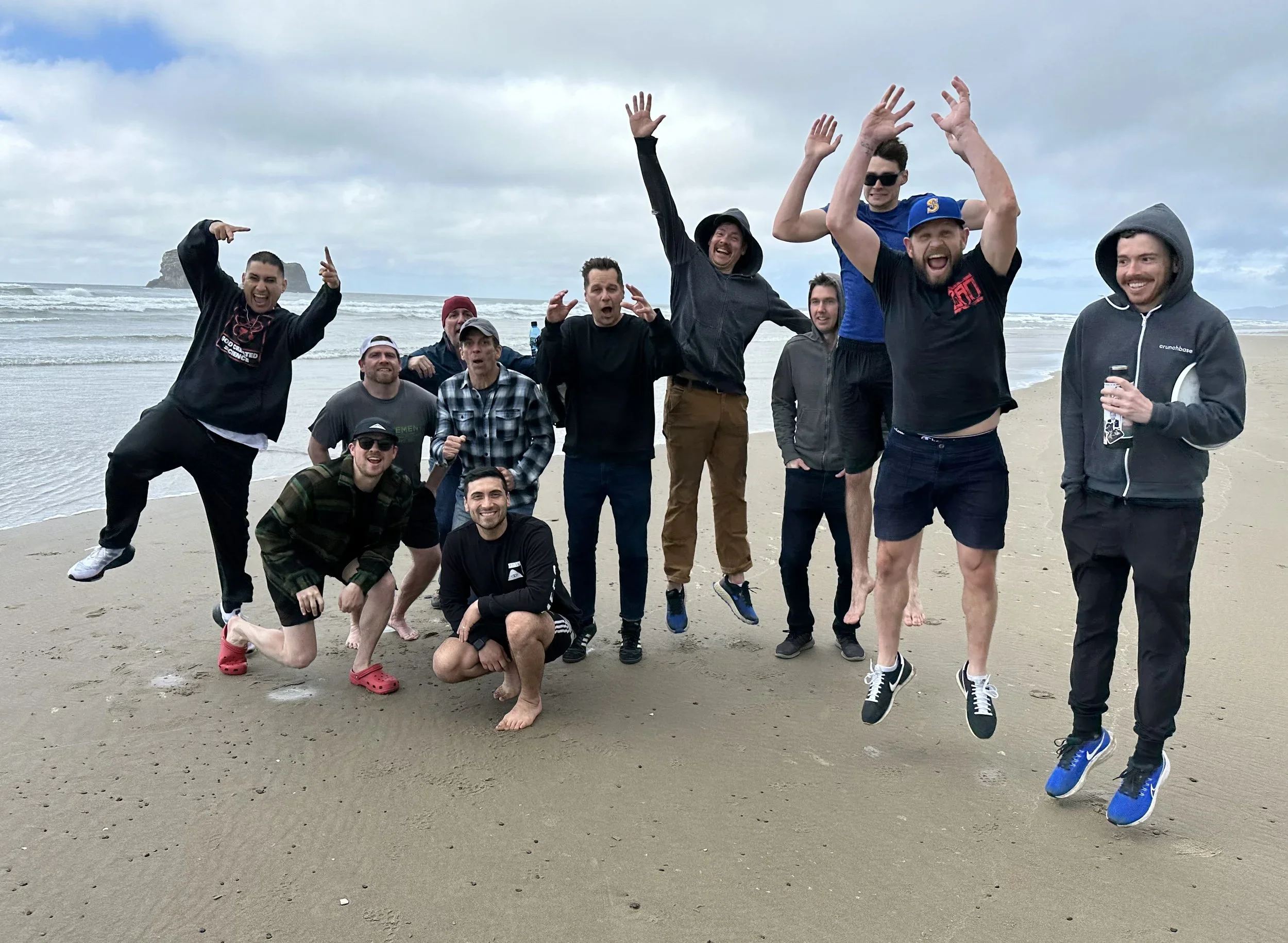 Group of eleven men on a beach, some jumping and smiling, with waves and cloudy sky in the background.