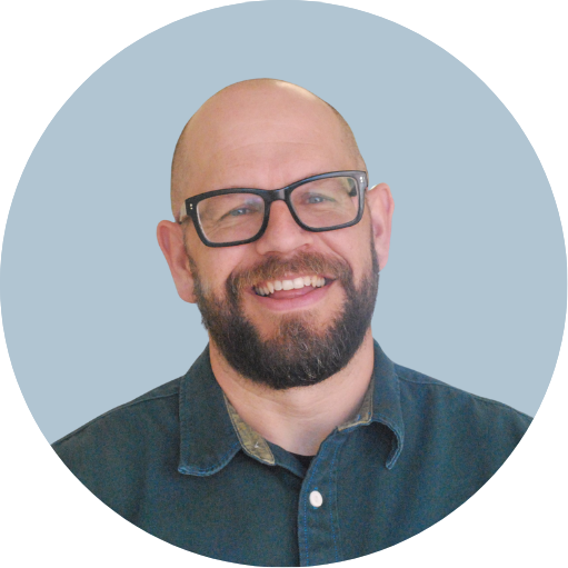 A smiling man with a bald head, beard, and glasses, wearing a blue denim shirt against a light blue circular background.