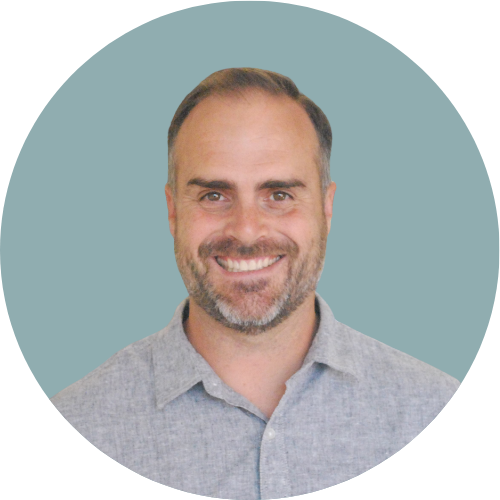A smiling man with short brown hair and a beard, wearing a light gray collared shirt, against a solid muted blue background.