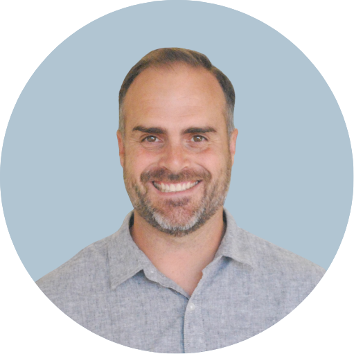 A smiling man with short brown hair and a beard, wearing a light gray collared shirt, against a solid muted blue background.