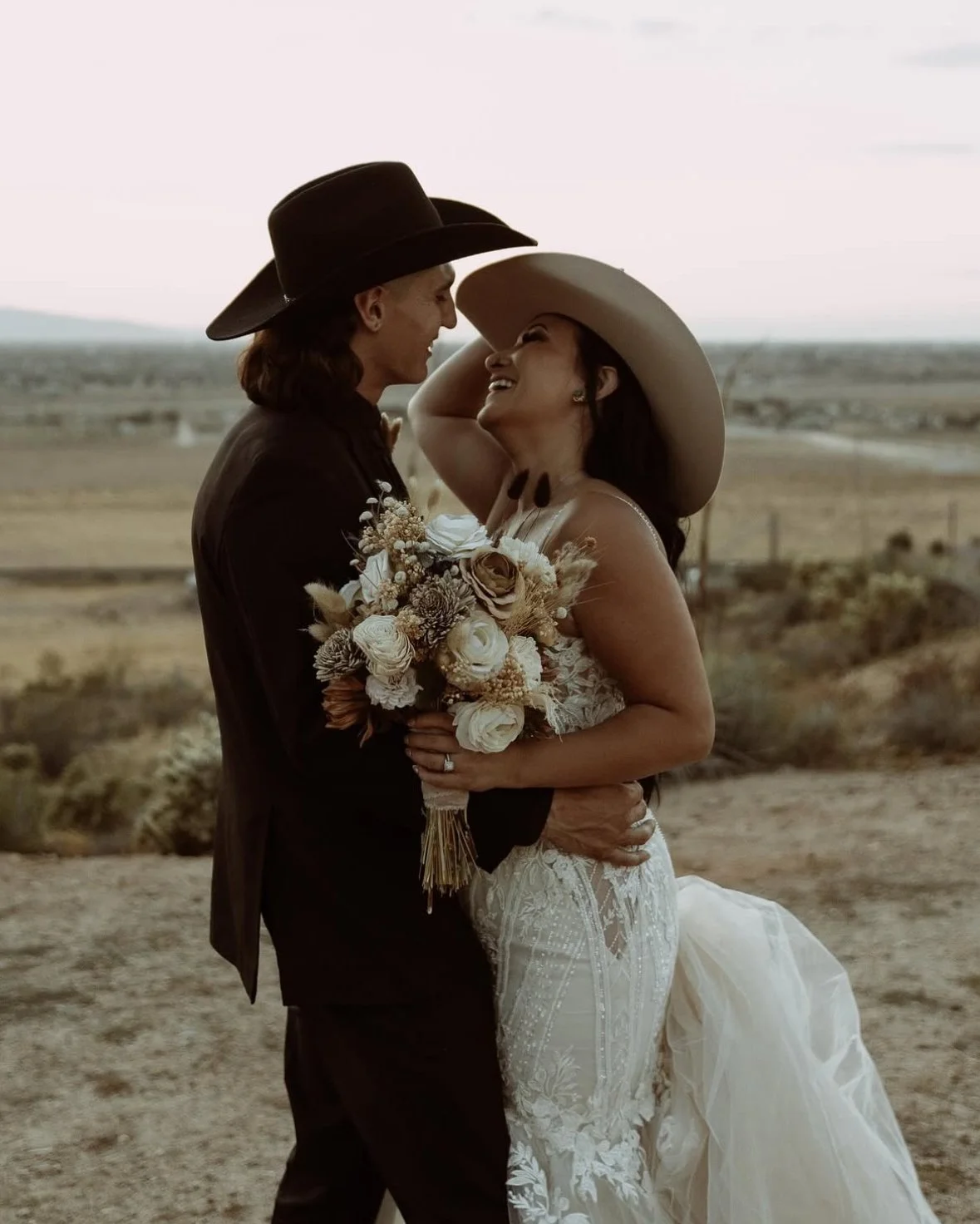 A couple in wedding attire, standing outdoors in a desert landscape, smiling and embracing each other while wearing cowboy hats. The woman is holding a bouquet of flowers and wearing a white wedding dress, and the man is in a black suit.