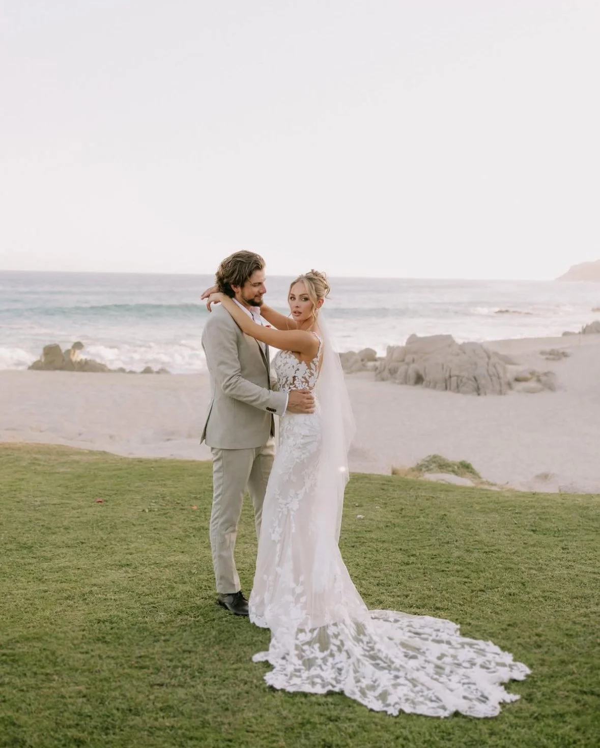 A bride and groom embrace on a grassy area near a beach, with rocky formations and ocean waves in the background, during their wedding photos.