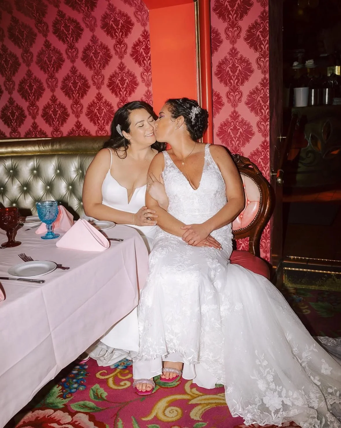 Two women in wedding dresses sharing a kiss at a decorated dining table with pink napkins, crystal glasses, and plates, sitting on a vintage-style chair in a room with red damask wallpaper.