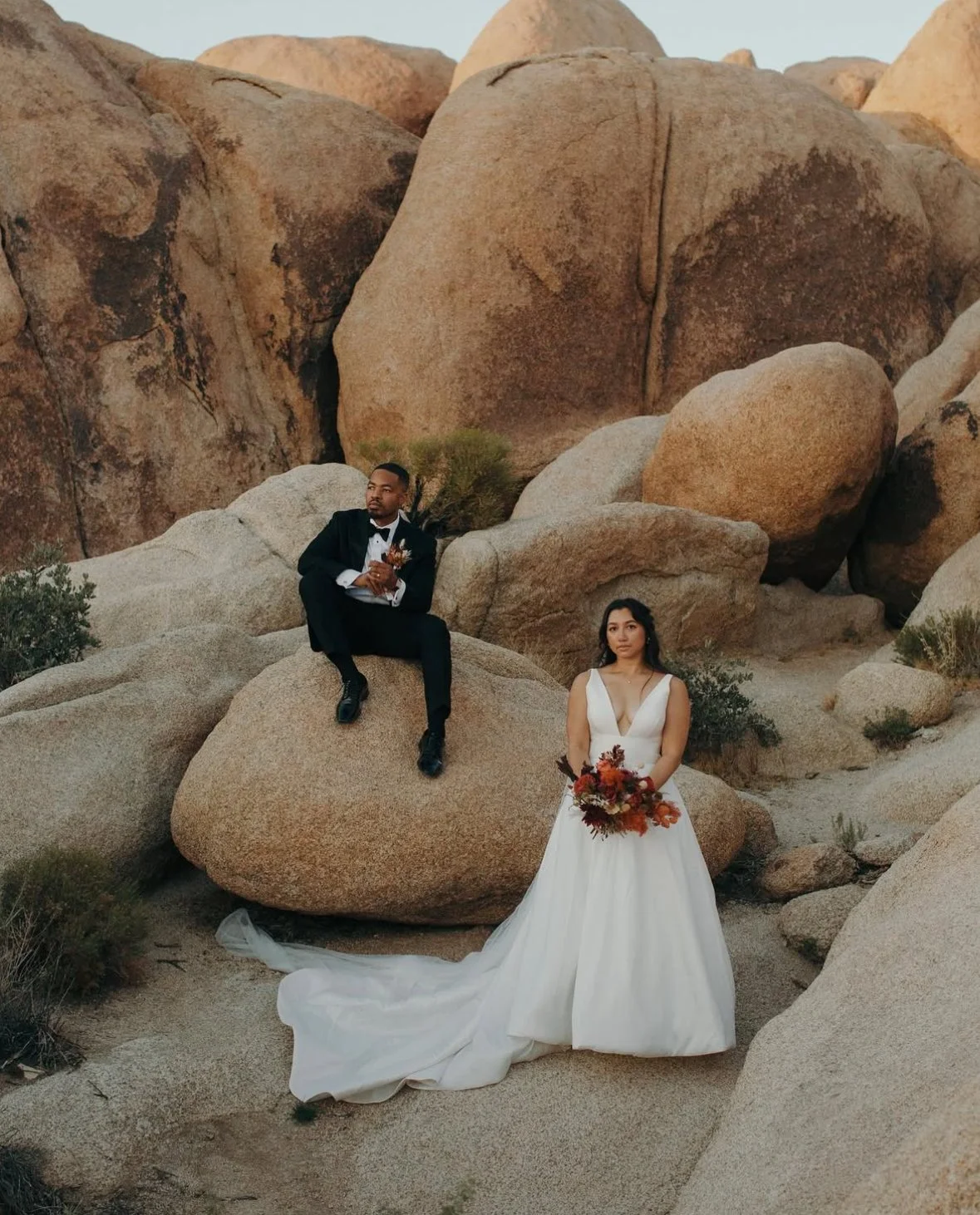 A bride in a white wedding dress holding a bouquet standing on sandy ground among rocks, with a groom dressed in a black tuxedo sitting on a large rock nearby in a desert-like landscape with large boulders.