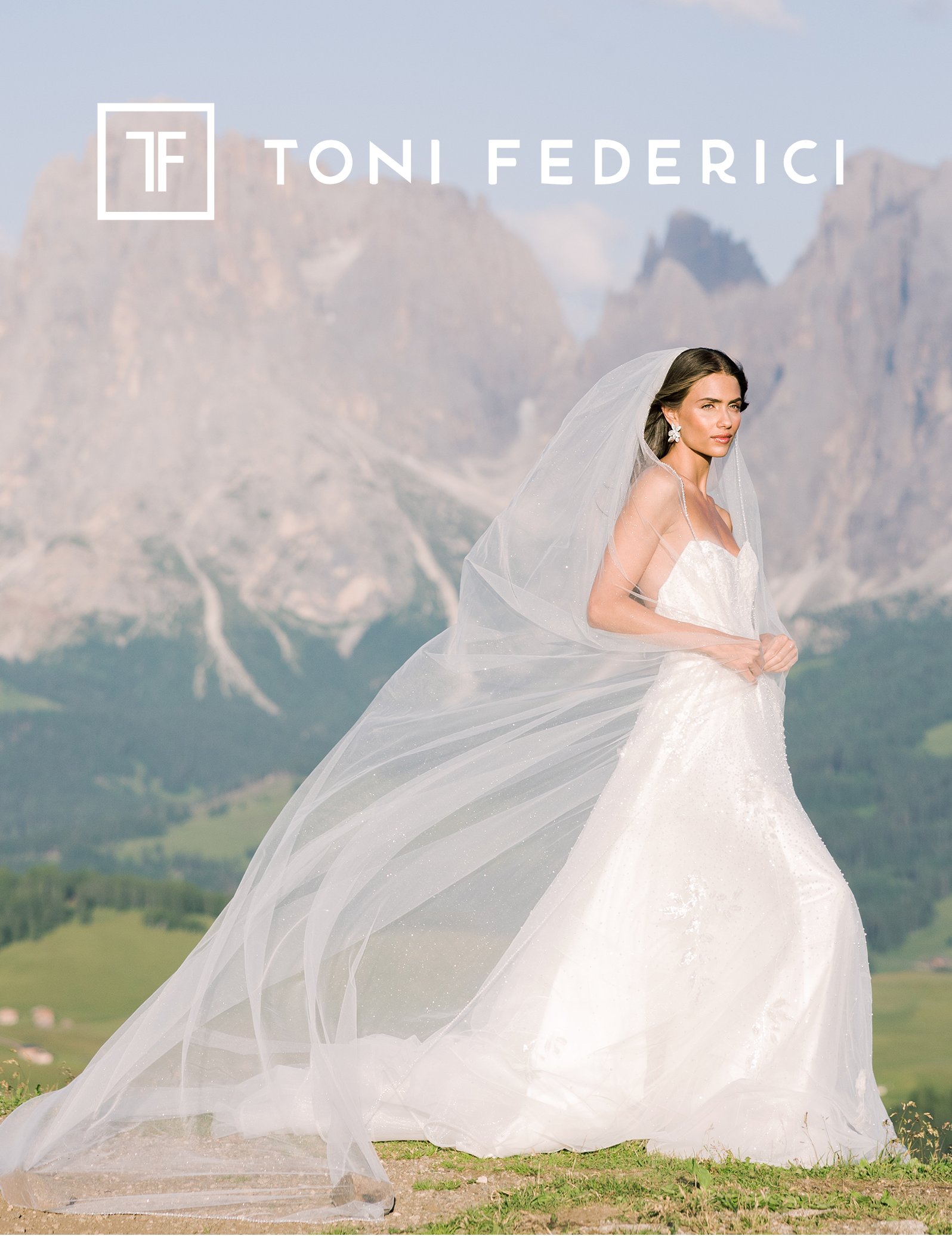 A woman in a wedding dress with a long, flowing veil standing outdoors with mountains in the background.
