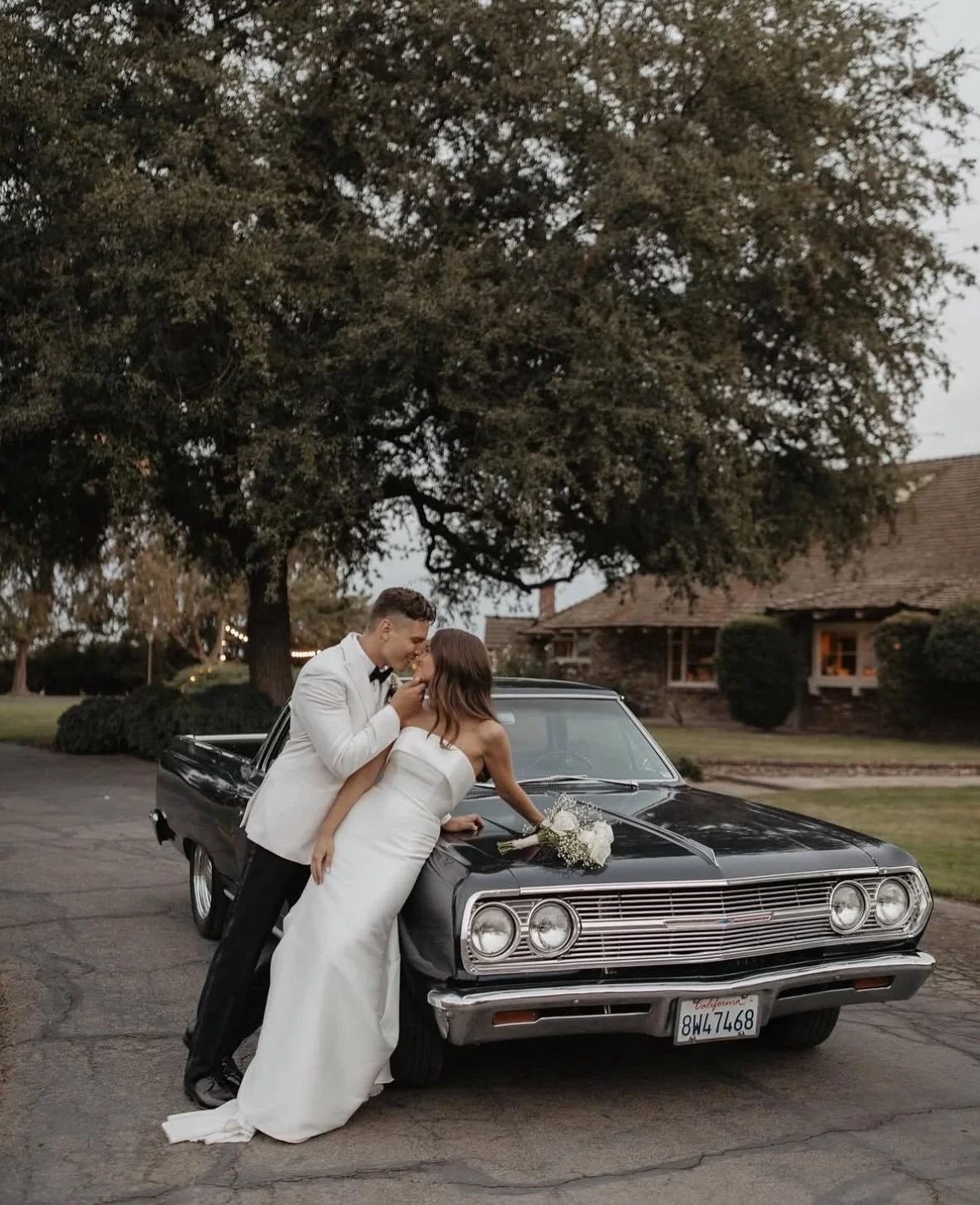 A newlywed couple sharing a kiss in front of a vintage black car with a bouquet of white flowers on the hood, outdoors with trees and a house in the background.