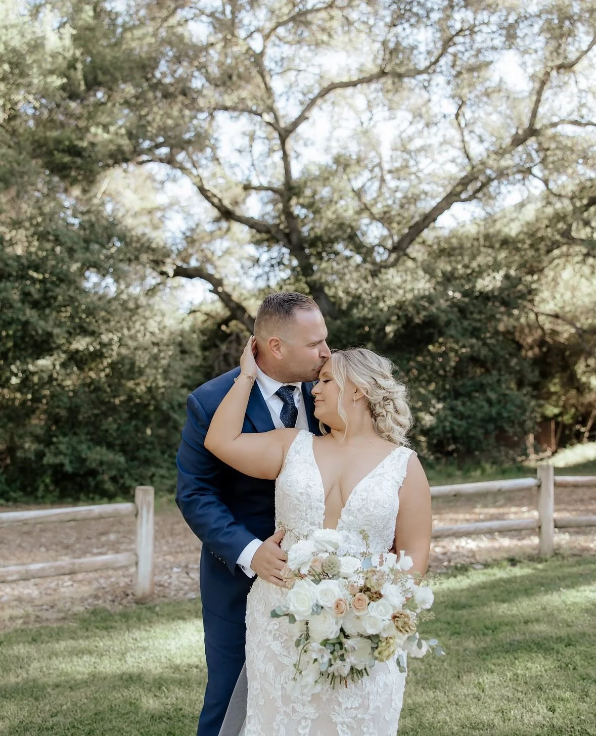 A newlywed couple embracing outdoors on a grassy area, with trees and a wooden fence in the background. The bride is holding a bouquet of white and blush flowers, and the groom is kissing her forehead.