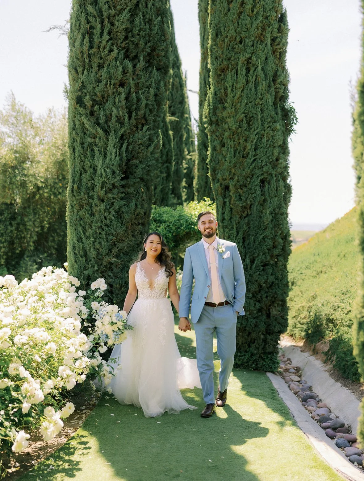 A bride and groom holding hands and smiling outdoors on their wedding day, walking along a grassy path lined with white flowers and tall green trees.
