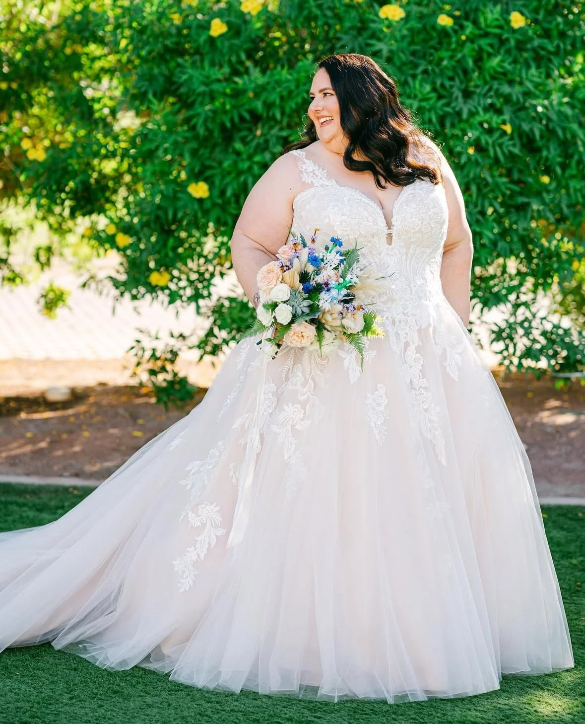 A woman in a white wedding dress holding a bouquet of flowers, standing outdoors near green bushes and trees.