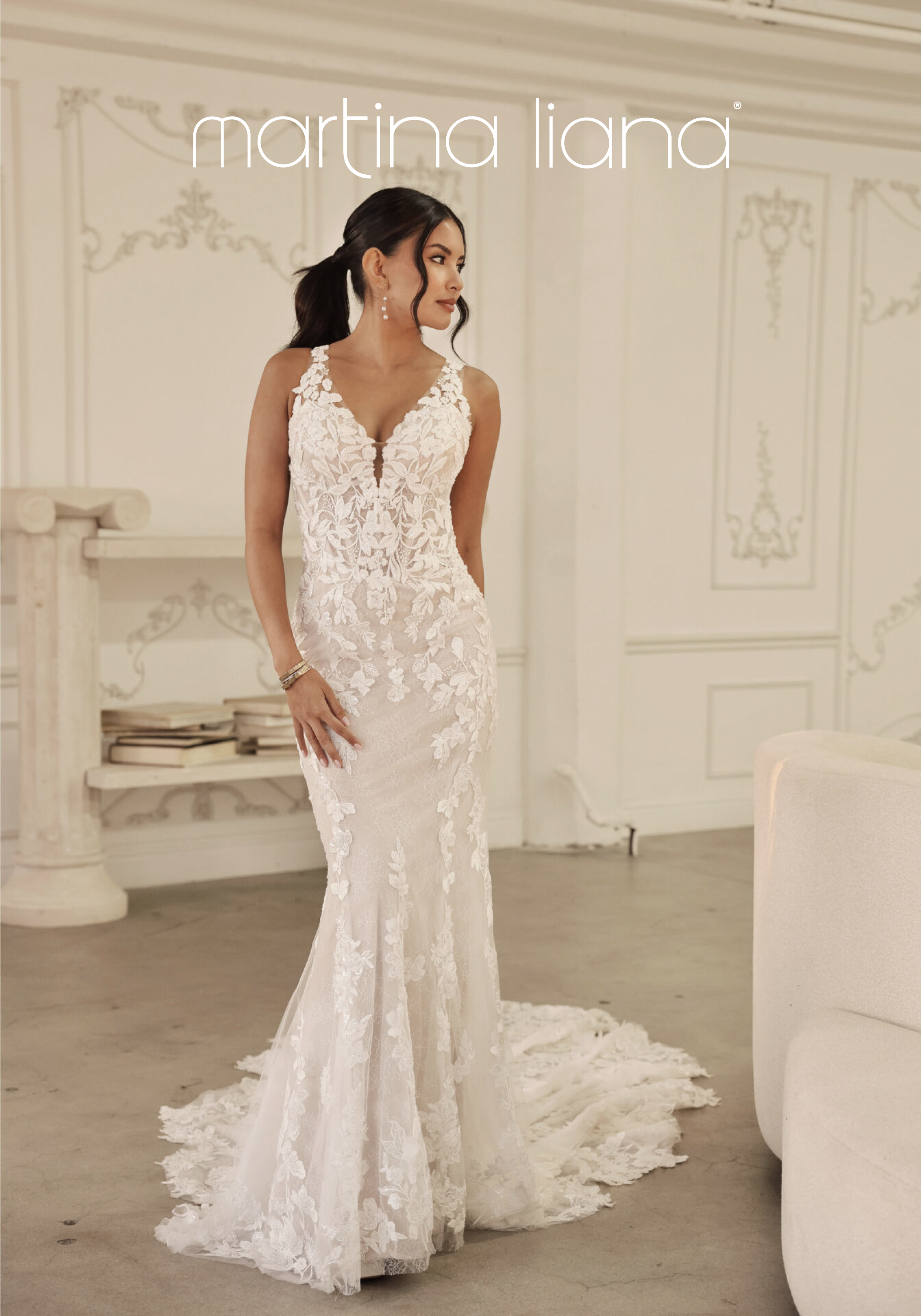 A woman wearing a white lace wedding dress with floral embroidery, standing in a decorated room with cream-colored walls.