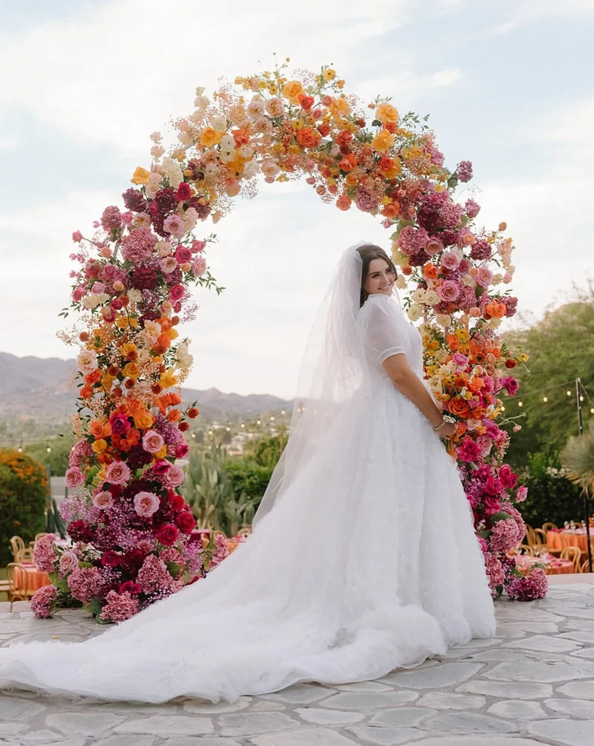 Bride in a white wedding gown and veil standing under a colorful flower arch outdoors, with mountains and a cloudy sky in the background.