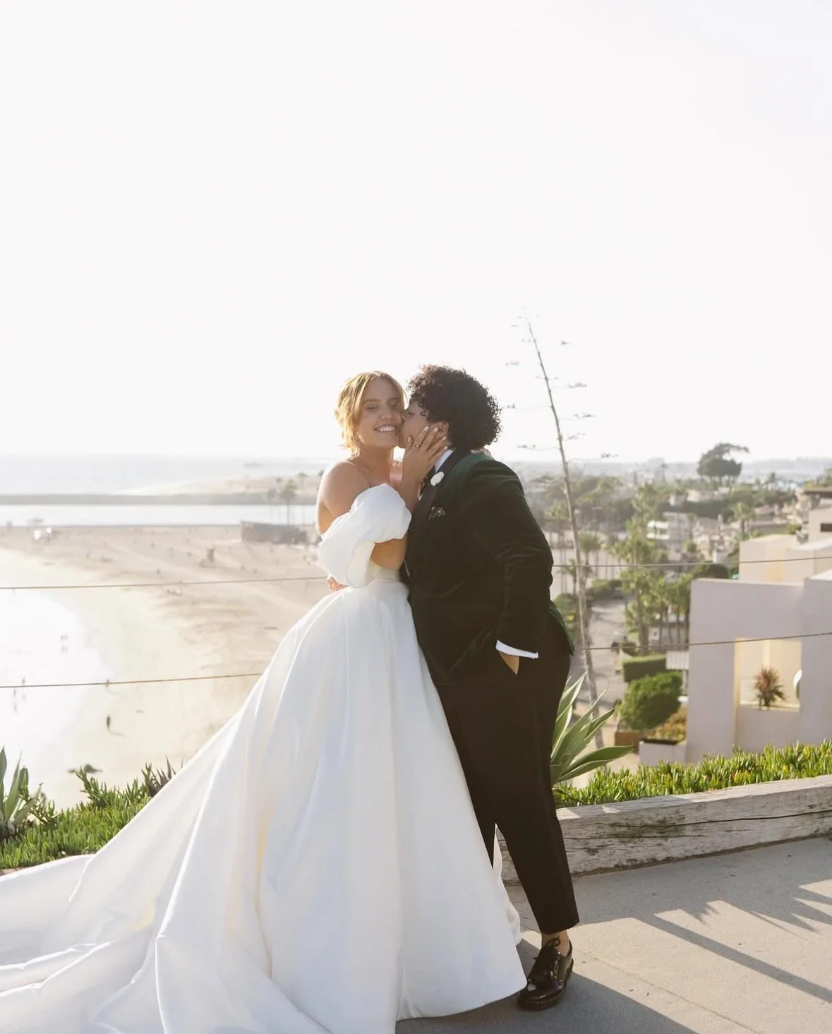 A bride in a white wedding dress and a groom in a black suit share a kiss and smile on a sunny day, with a beach and ocean in the background.