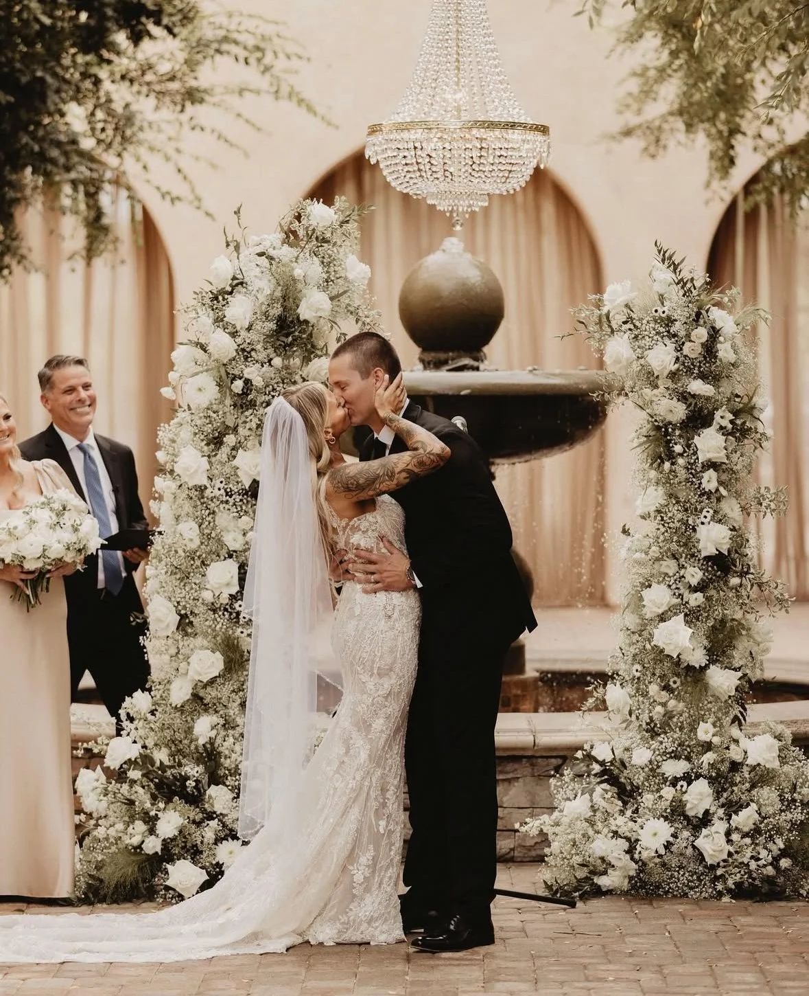 A bride and groom sharing a kiss during their wedding ceremony, standing in front of a backdrop with floral arrangements and a chandelier.