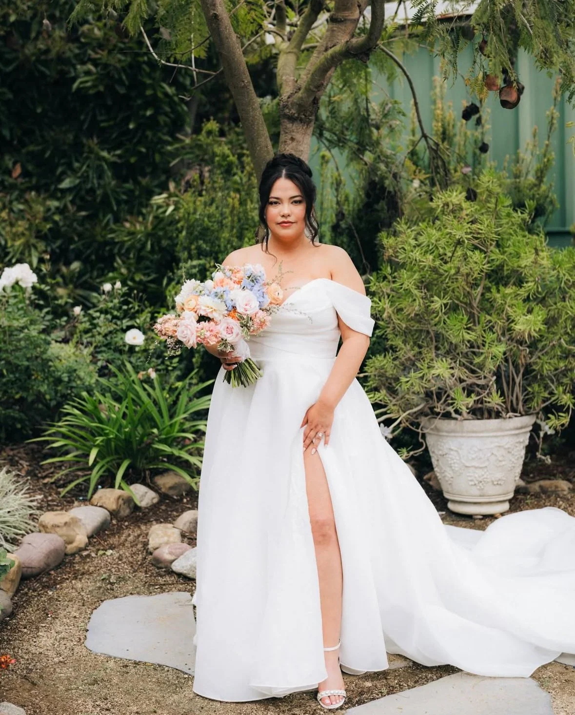 A woman in a white wedding dress holding a bouquet of pastel-colored flowers, standing outdoors in a lush garden with trees and plants.