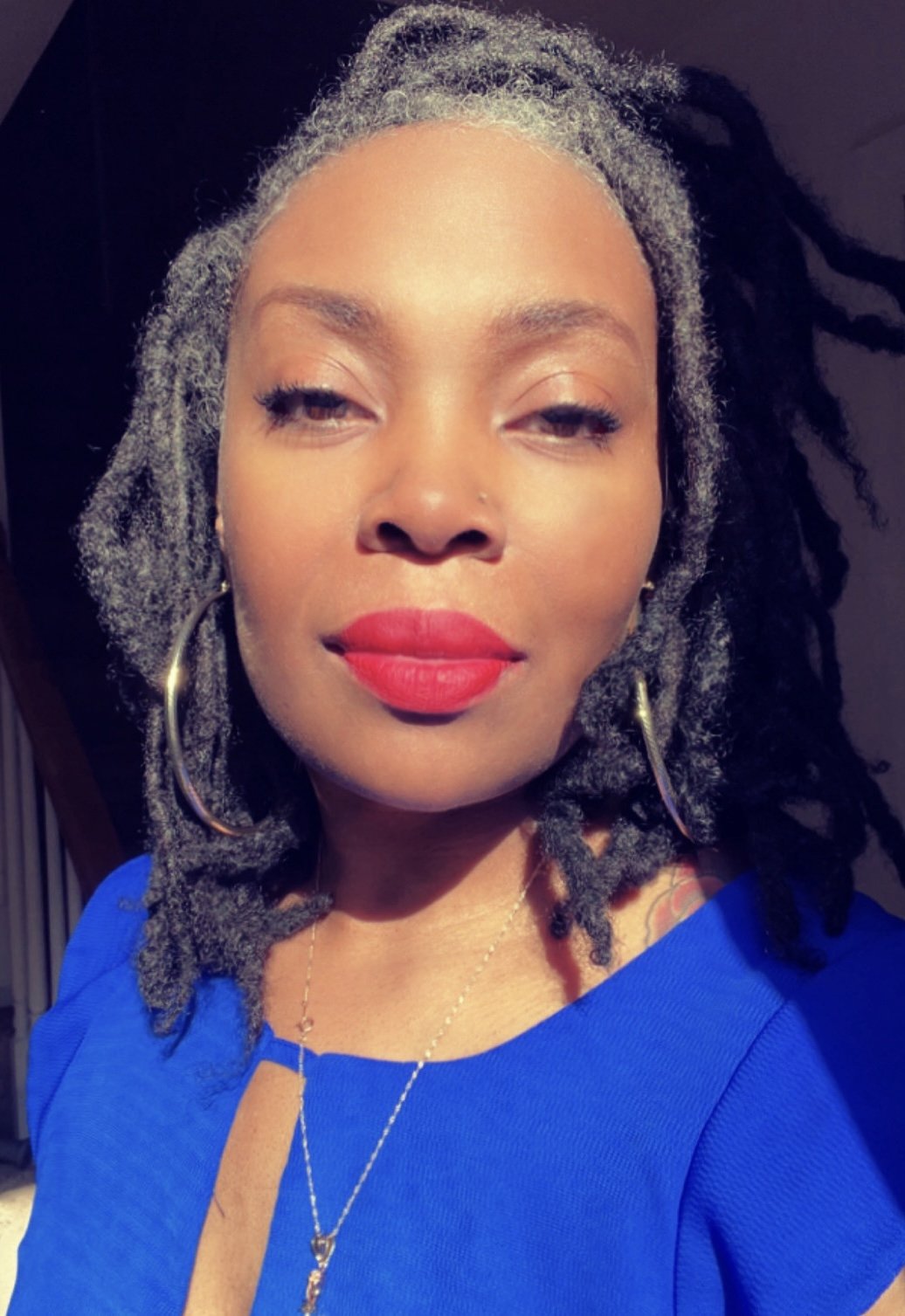 A woman with gray dreadlocks, wearing a blue top, gold hoop earrings, a gold necklace, and bright red lipstick taking a selfie with a relaxed expression.