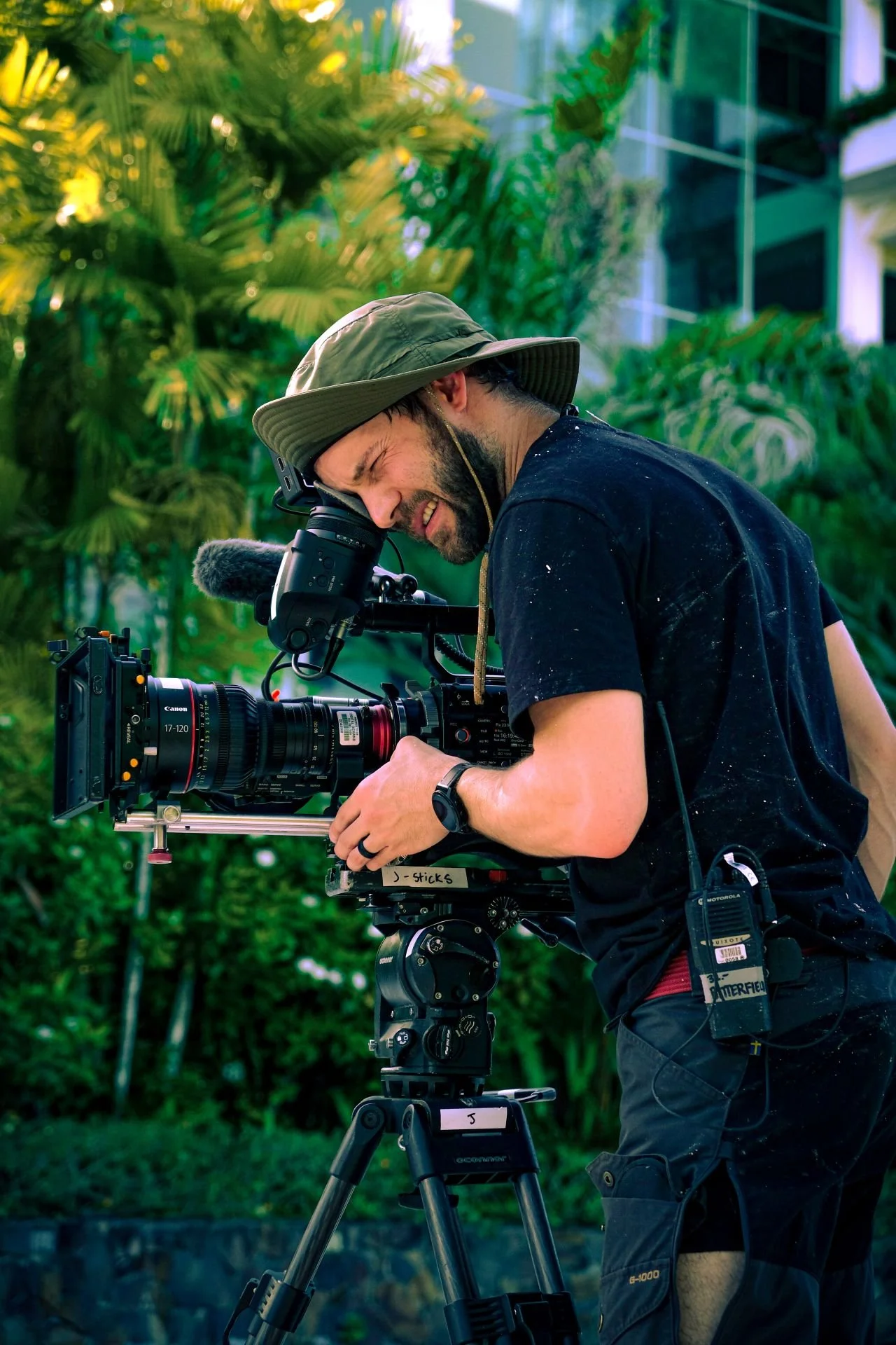 A man operating a professional film camera outdoors with greenery in the background.