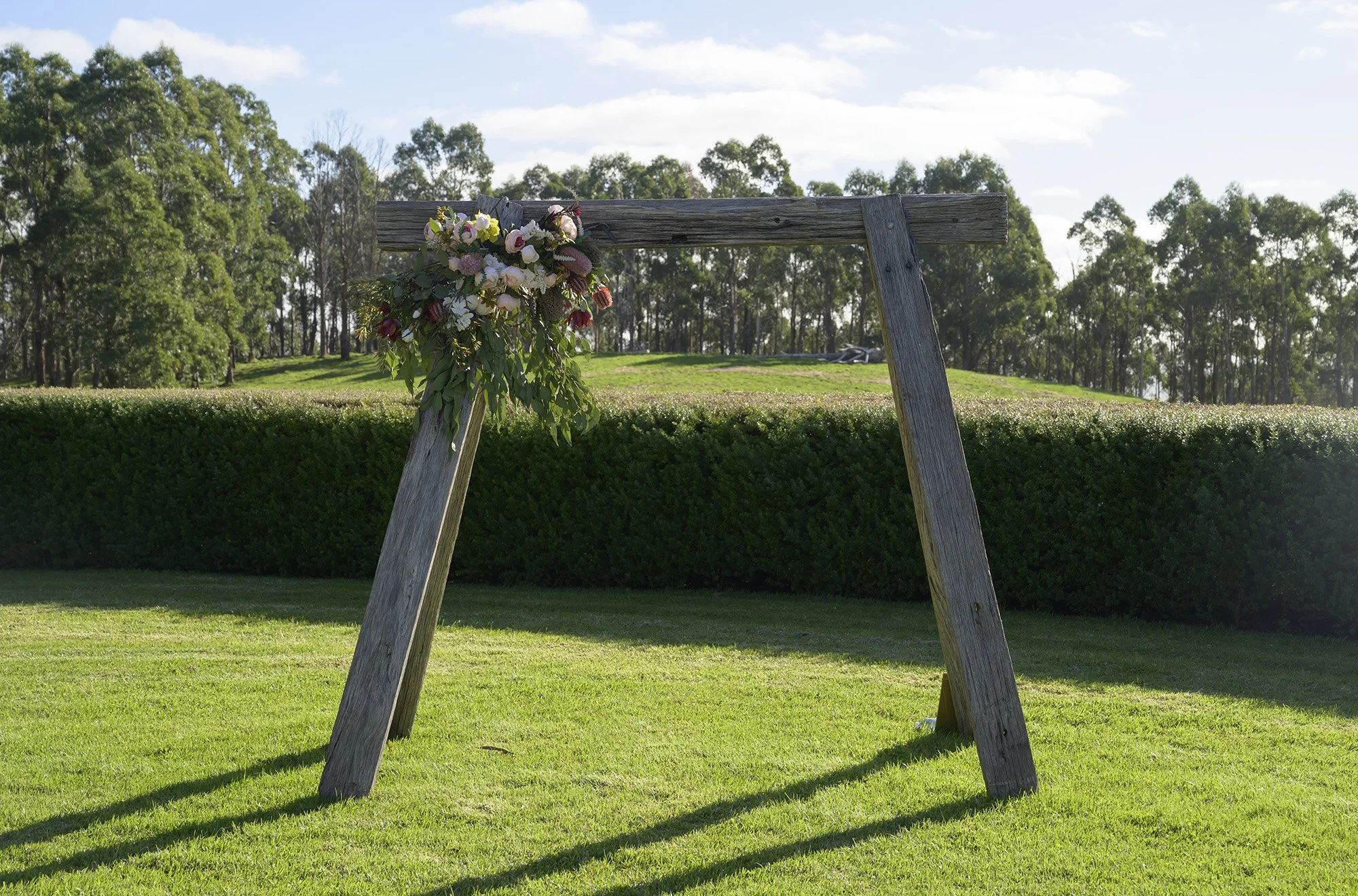 A wooden arch with a floral arrangement on the top left corner, set on a grassy outdoor area with trees in the background.
