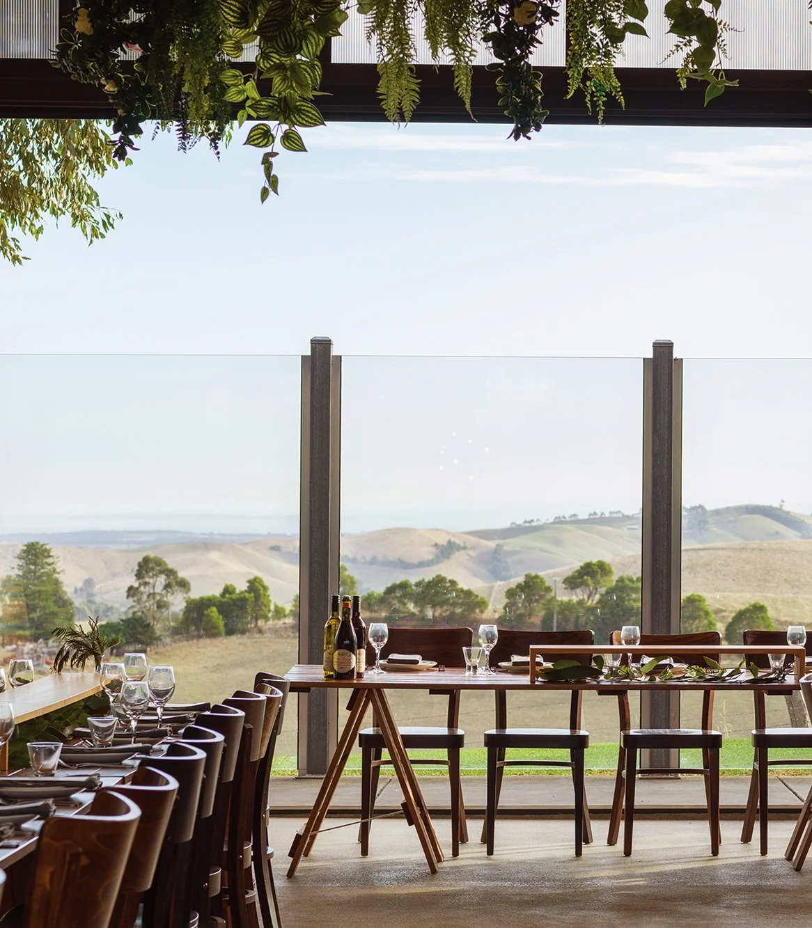 Indoor dining area with a view of South Gippsland rolling hills and trees through large glass windows, set with chairs, tables, wine bottles, and glassware for the perfect wedding.