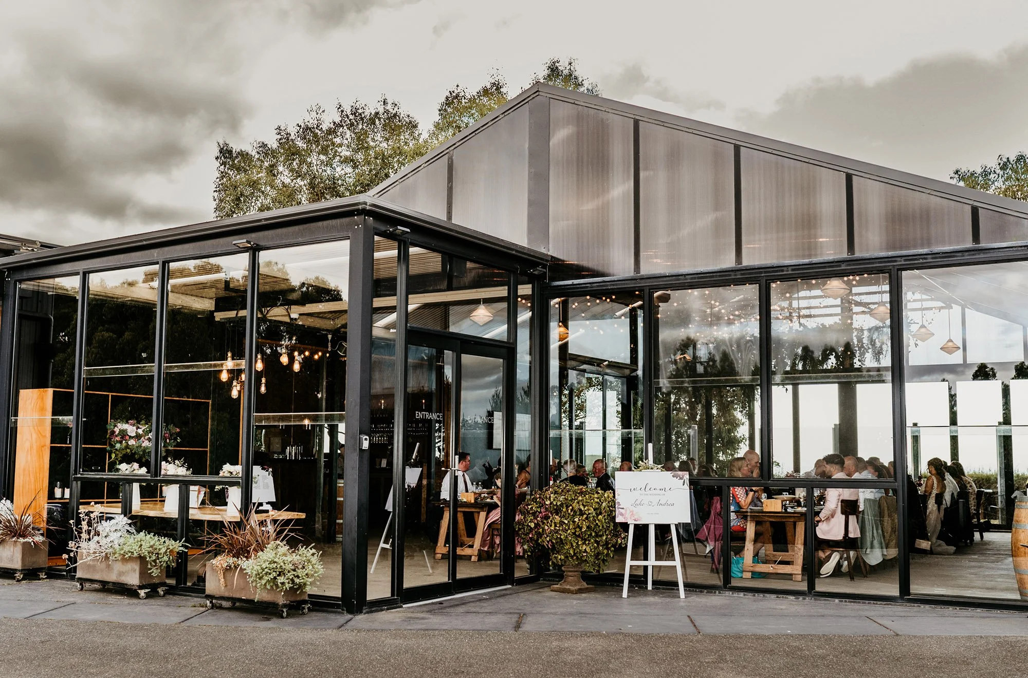 Glass-walled restaurant or event space with people dining inside, decorated with string lights and plants, during cloudy weather