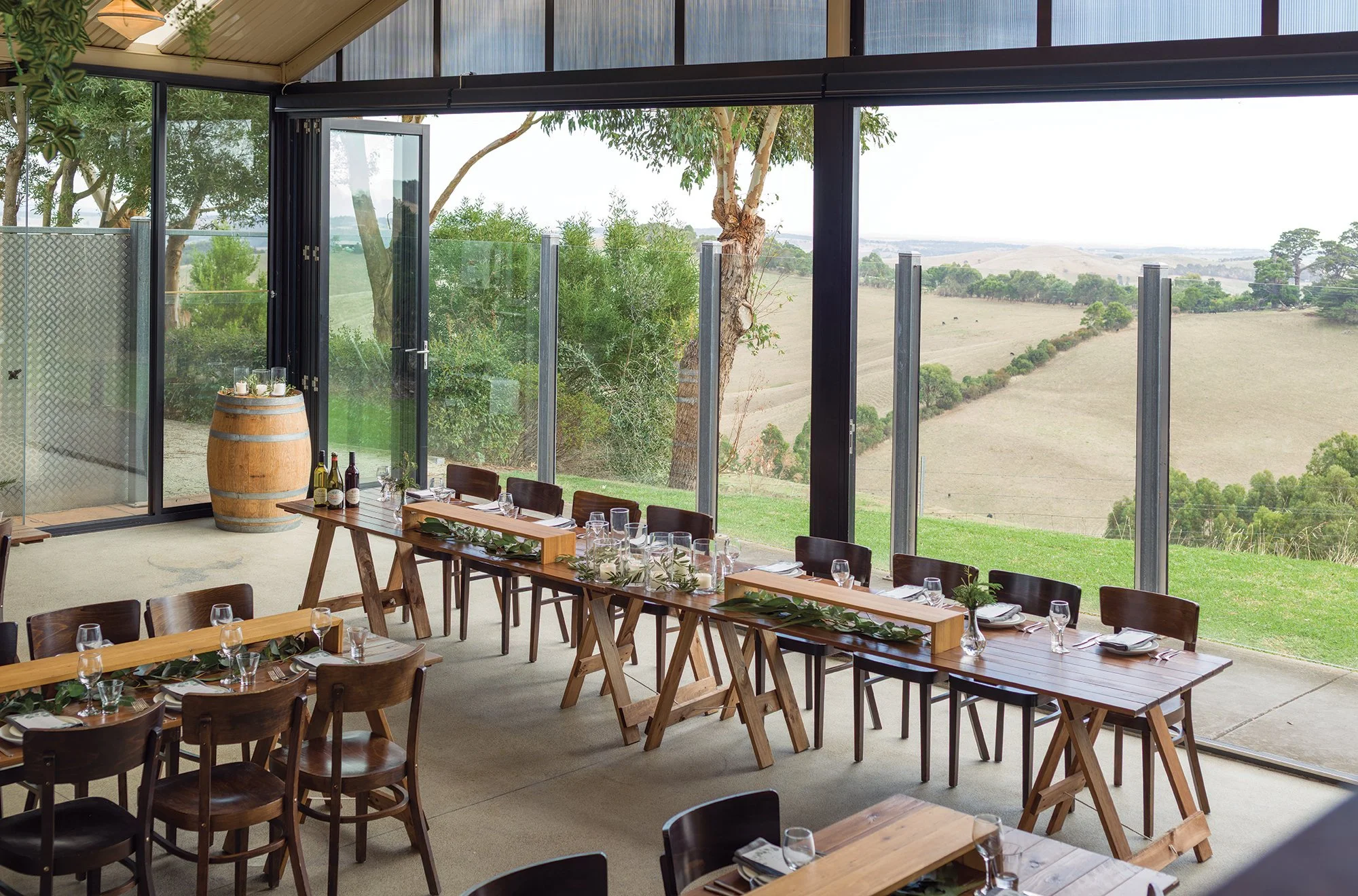 Elegant dining area with wooden tables and chairs set for a meal, decorated with greenery, inside a room with large glass windows showcasing a scenic rural landscape with trees, rolling hills, and open fields.