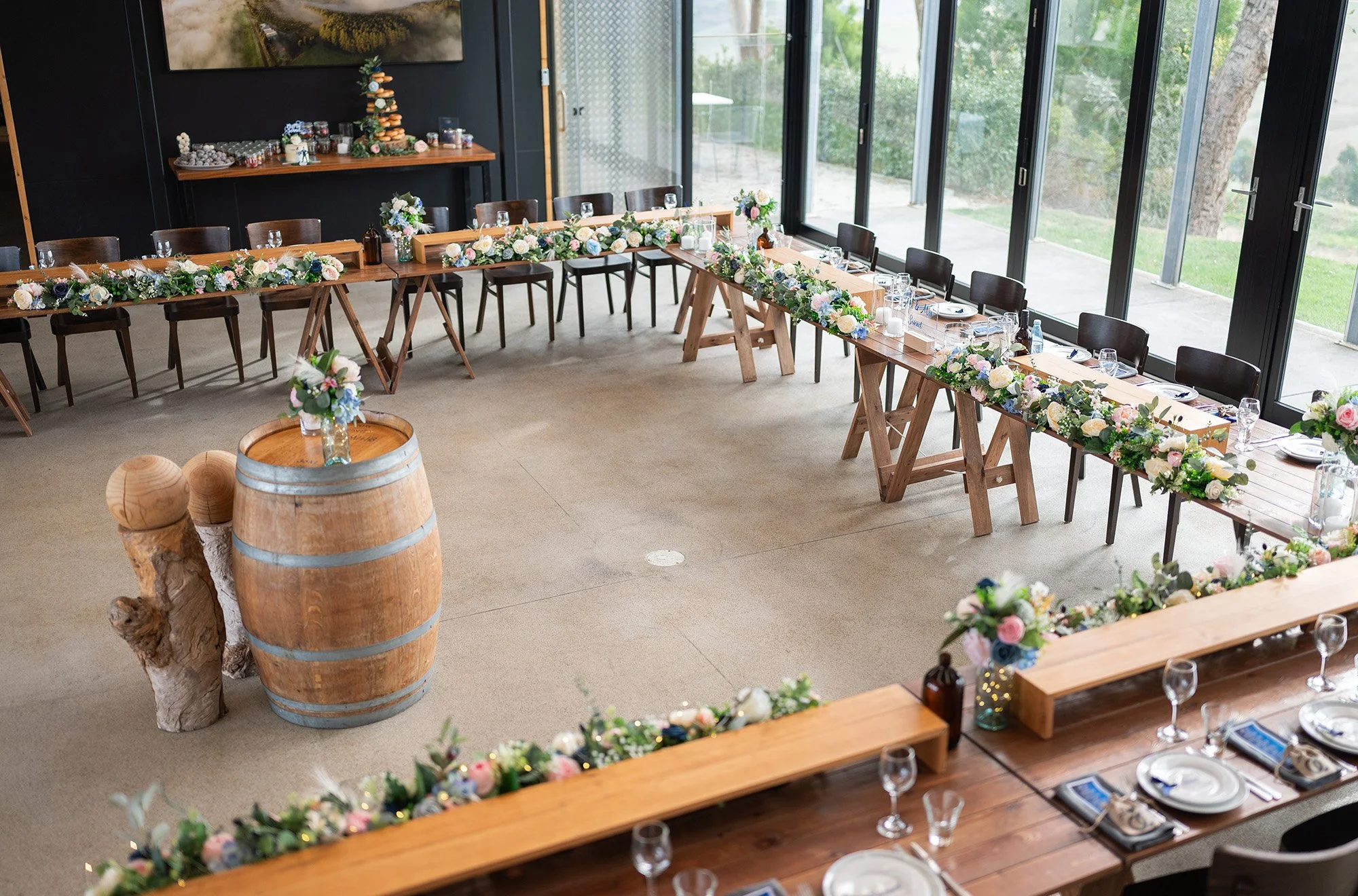 A decorated indoor dining area with a U-shaped table setup for a celebration, featuring floral garlands, flower arrangements, and table settings, with a barrel and carved wood piece in the center and large windows on one side letting in natural light
