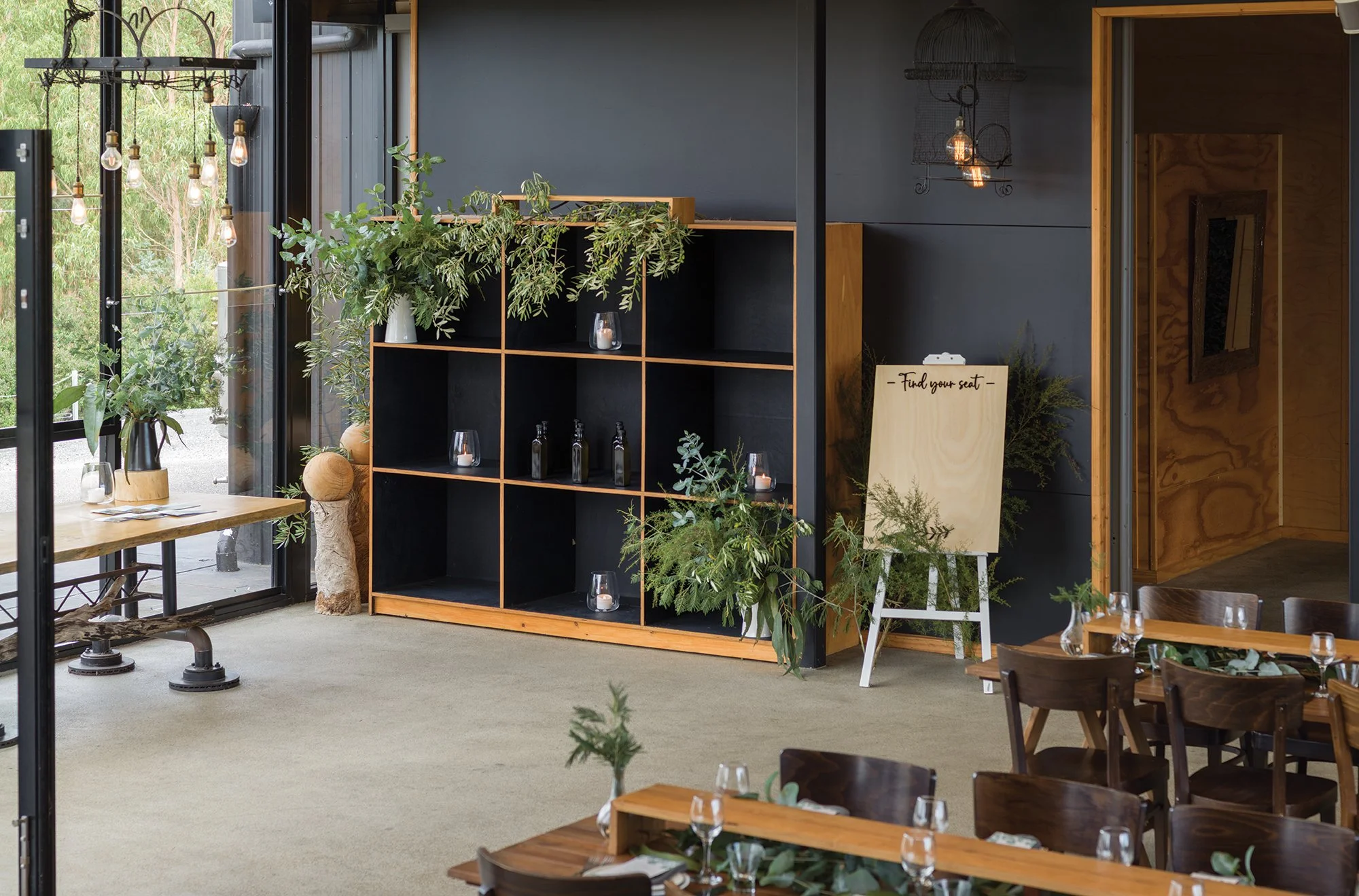 Interior of a modern, stylish restaurant with wooden tables and chairs, decorated with leafy greenery, glassware, and candles. A large black shelving unit displays plants, candles, and bottles. There is a sign on a white easel that says 'Find your se