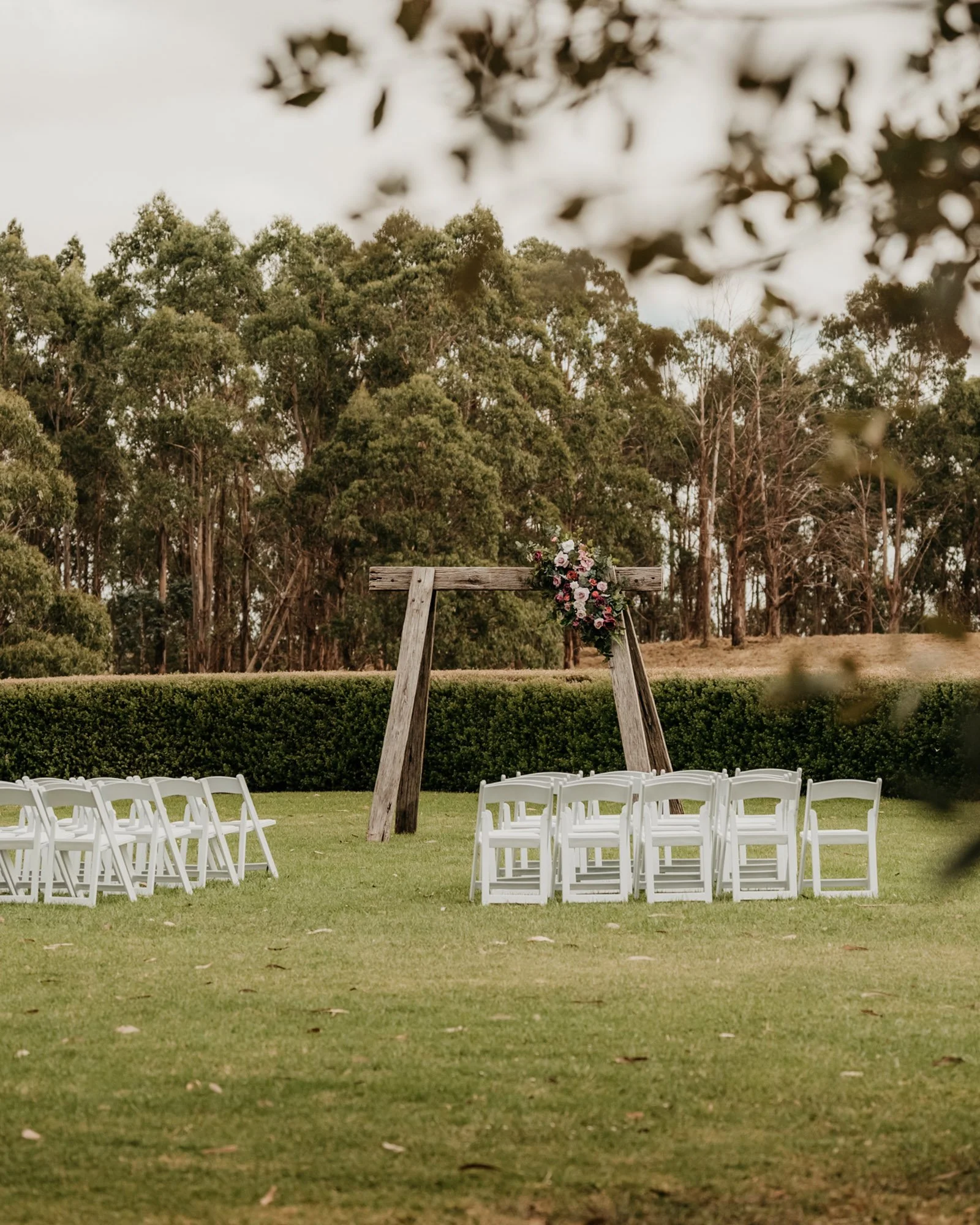 Outdoor wedding setup with a wooden arch decorated with flowers, surrounded by white chairs on a grassy field with trees in the background.