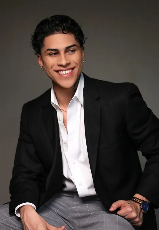 Young man with dark curly hair, smiling, wearing a black blazer over a white shirt, seated against a neutral background.