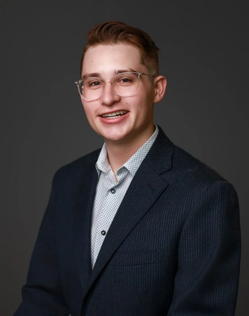 A young man with short brown hair, glasses, and light skin wearing a navy blazer and a white patterned shirt, smiling at the camera against a dark gray background.