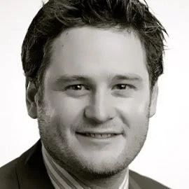 A black-and-white headshot of a young man with dark hair, smiling, wearing a suit and tie.
