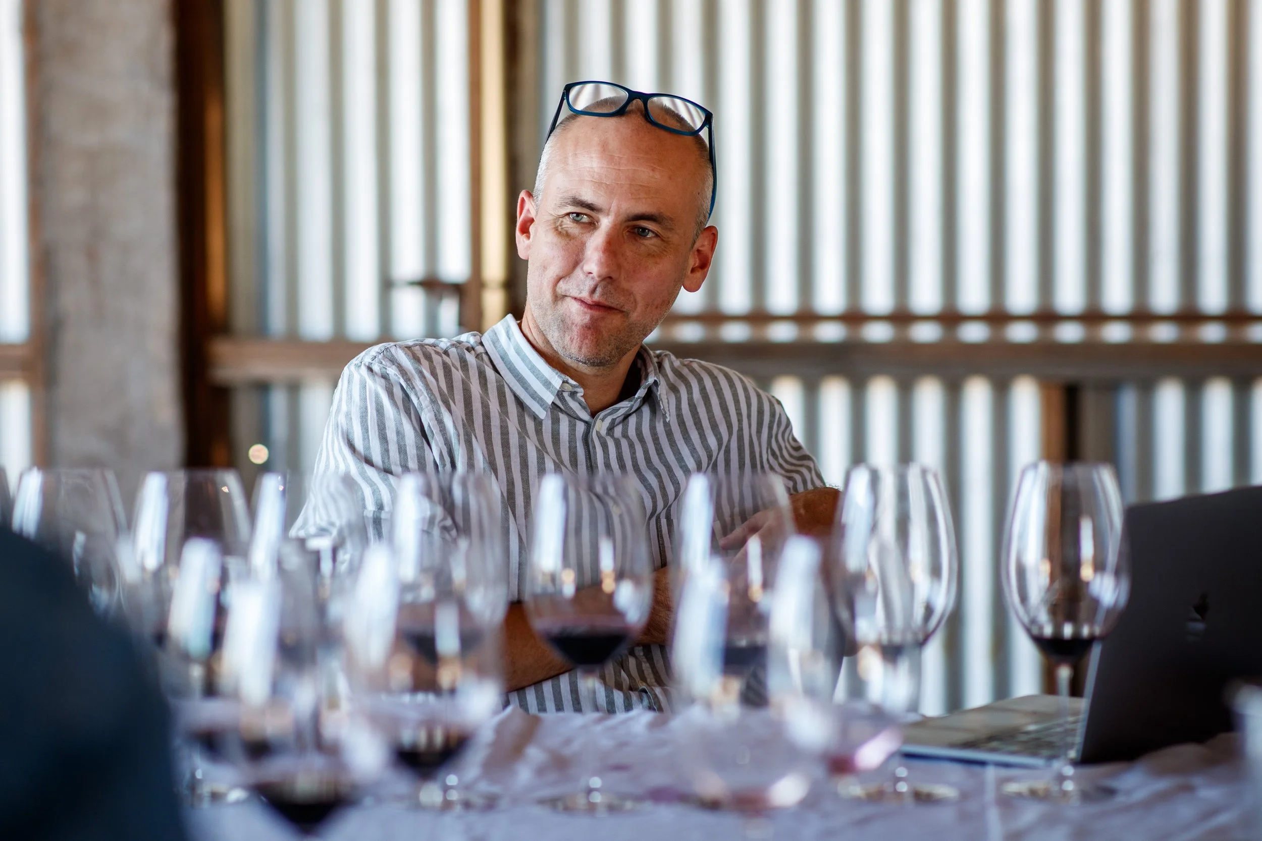 A man sitting at a table with multiple wine glasses, wearing a striped shirt, with glasses resting on his head, in a setting with wooden paneling and a blurred background.